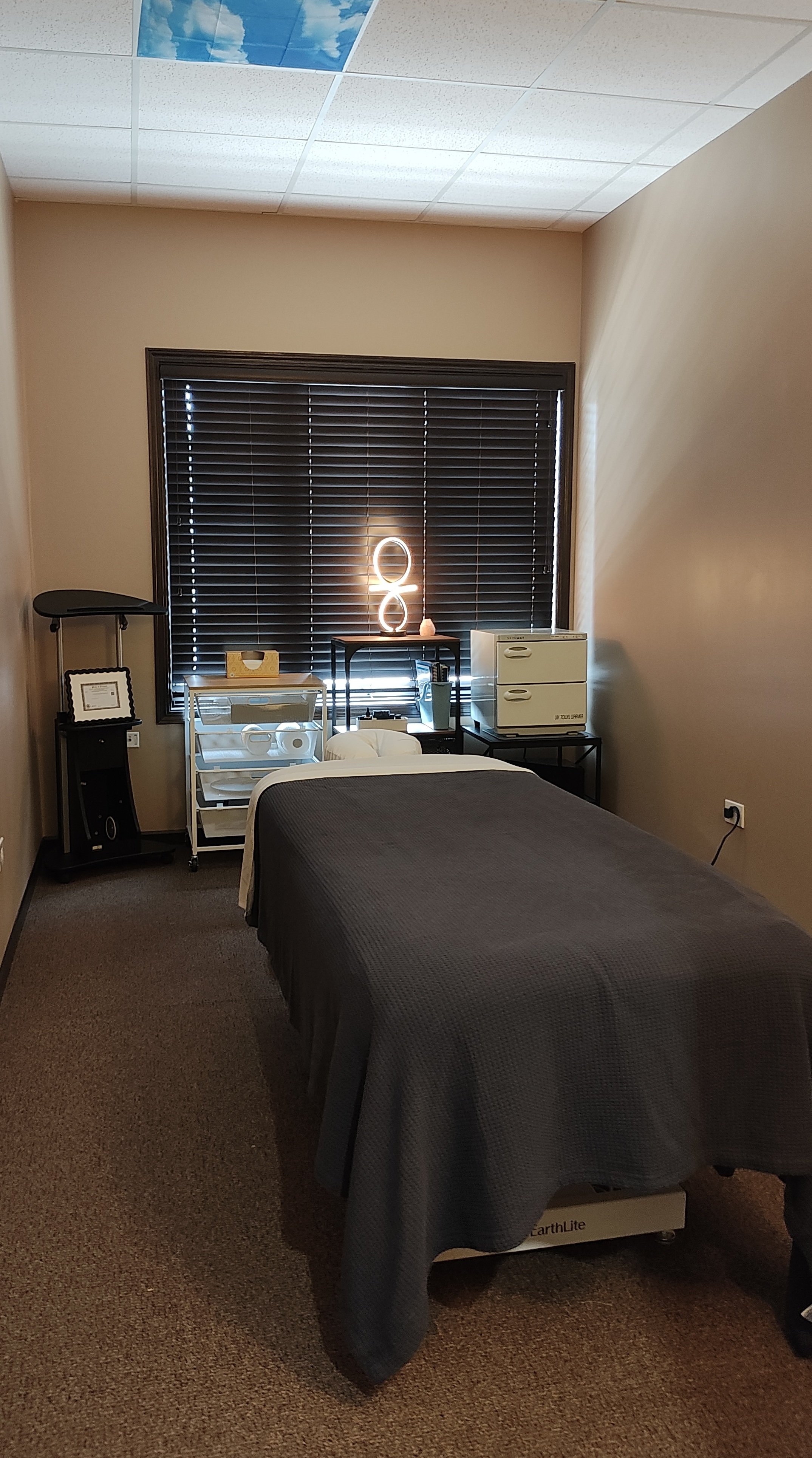 A massage or therapy room with a woman lying face down on a massage table. The room has beige walls, a black window with closed blinds, and a ceiling with ceiling tiles. There is a black side table with a decorative lamp, a white cart holding towels and a small decorative item, and medical or therapy equipment including a small white cabinet and a black oxygen concentrator.