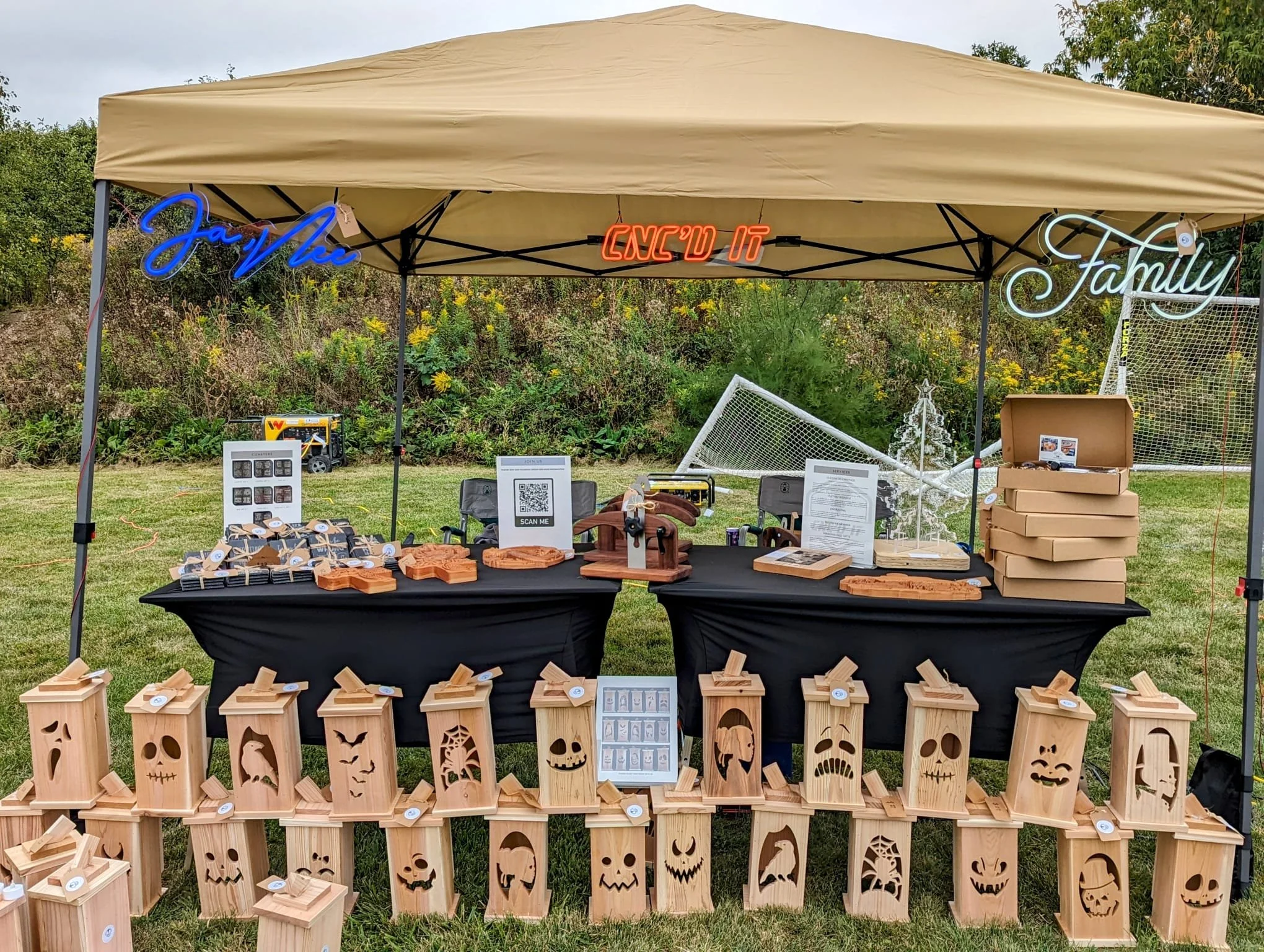 Craft fair booth under a tan canopy displaying handmade wooden lanterns with carved patterns, small wooden items for sale, and decorative signs in neon lights."