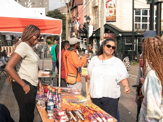 Two women laughing and talking at an outdoor street market stall with snacks and beverages, surrounded by pedestrians and colorful buildings.