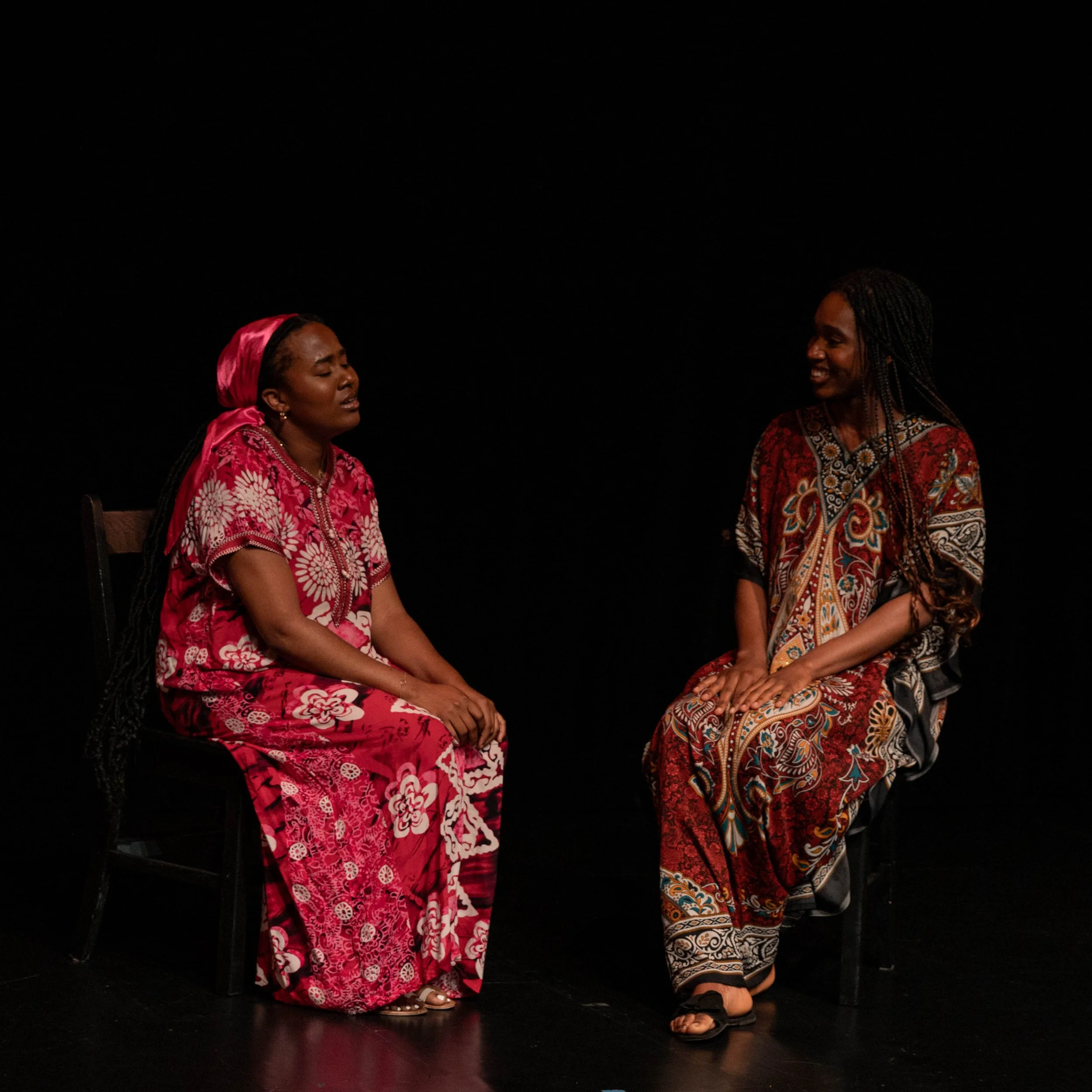 Two women sit on chairs facing each other against a black background, engaged in conversation. The woman on the left wears a pink and white floral dress and matching headscarf, with a serious expression. The woman on the right wears a multicolored, patterned dress and is smiling warmly.