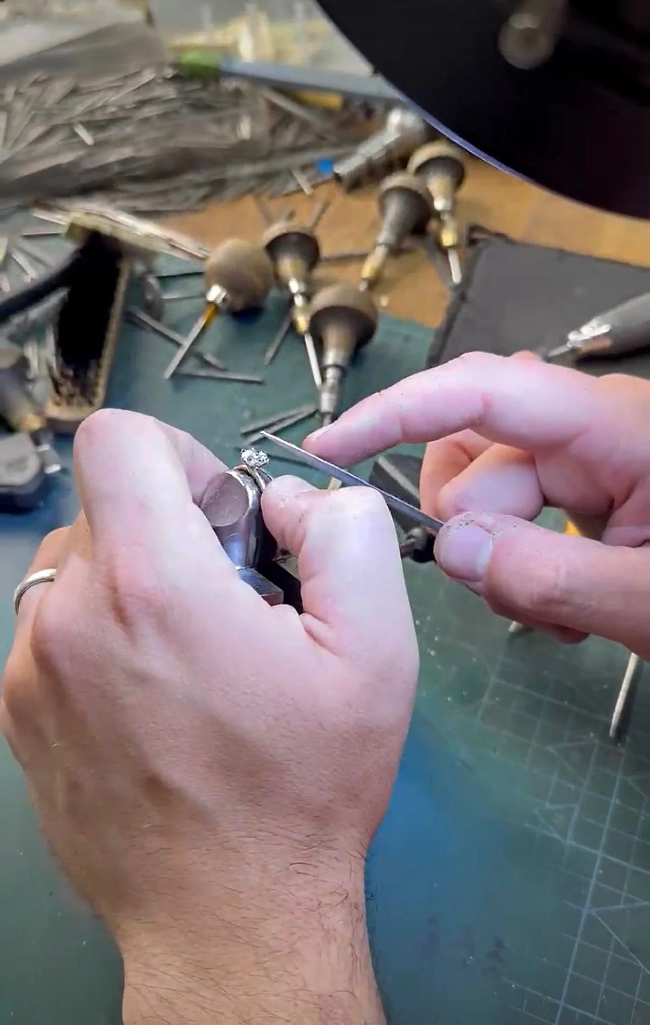 A jeweler is working on a ring with a diamond, holding a small tool. Various polishing and engraving tools are on the workbench in the background.