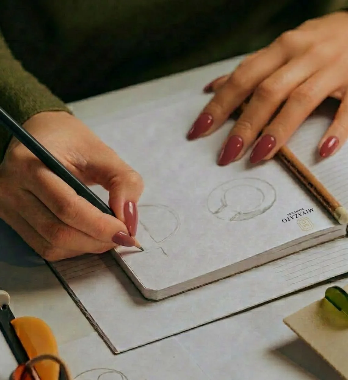 Person drawing on a spiral notebook with a black pen, holding the paper steady with their other hand which has painted nails. The notebook has the label "MIYAZATO" on the cover.