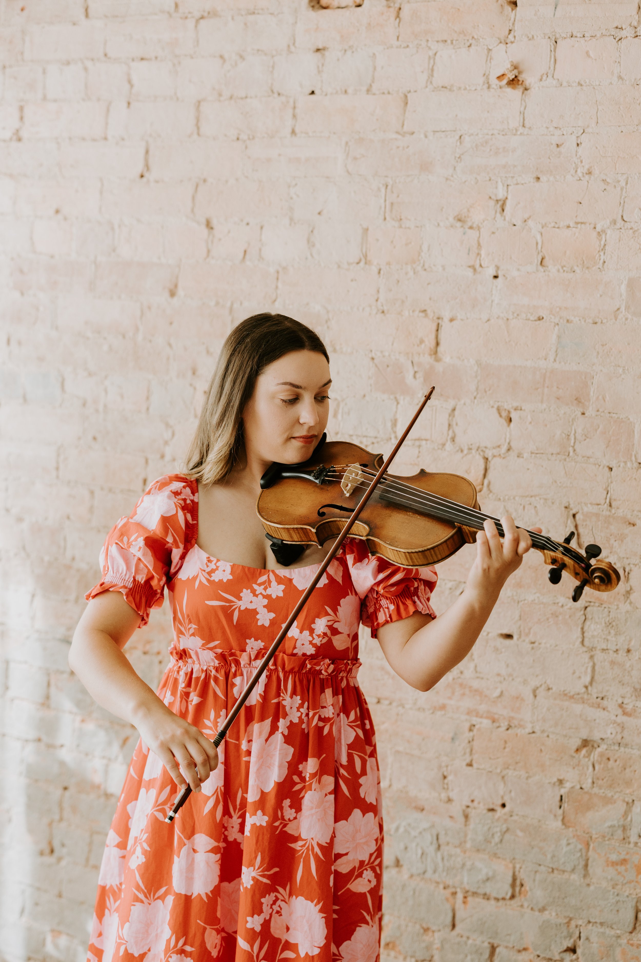 A woman in a red floral dress playing the violin against a beige brick wall.