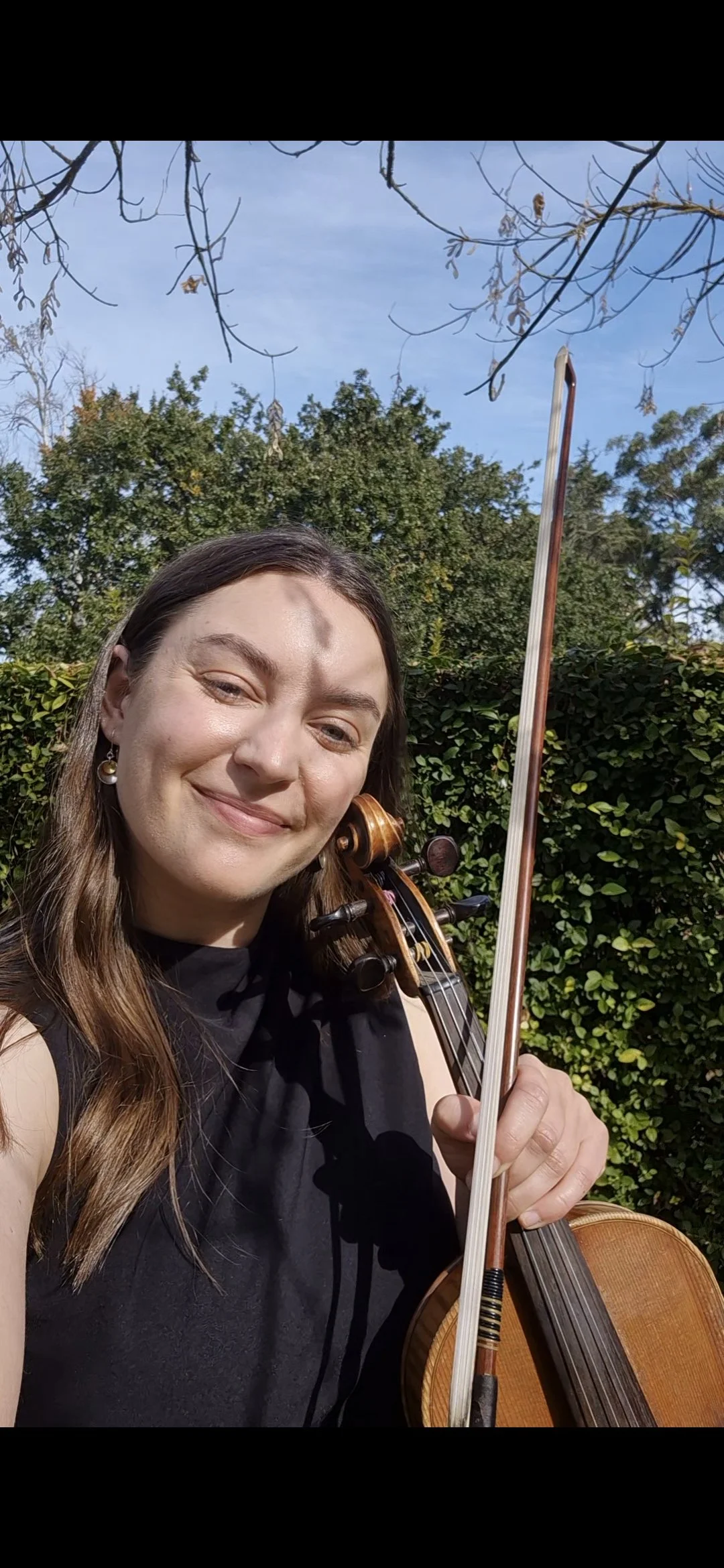 A woman with long brown hair holding a violin and bow, smiling outdoors with green bushes and trees under a blue sky.