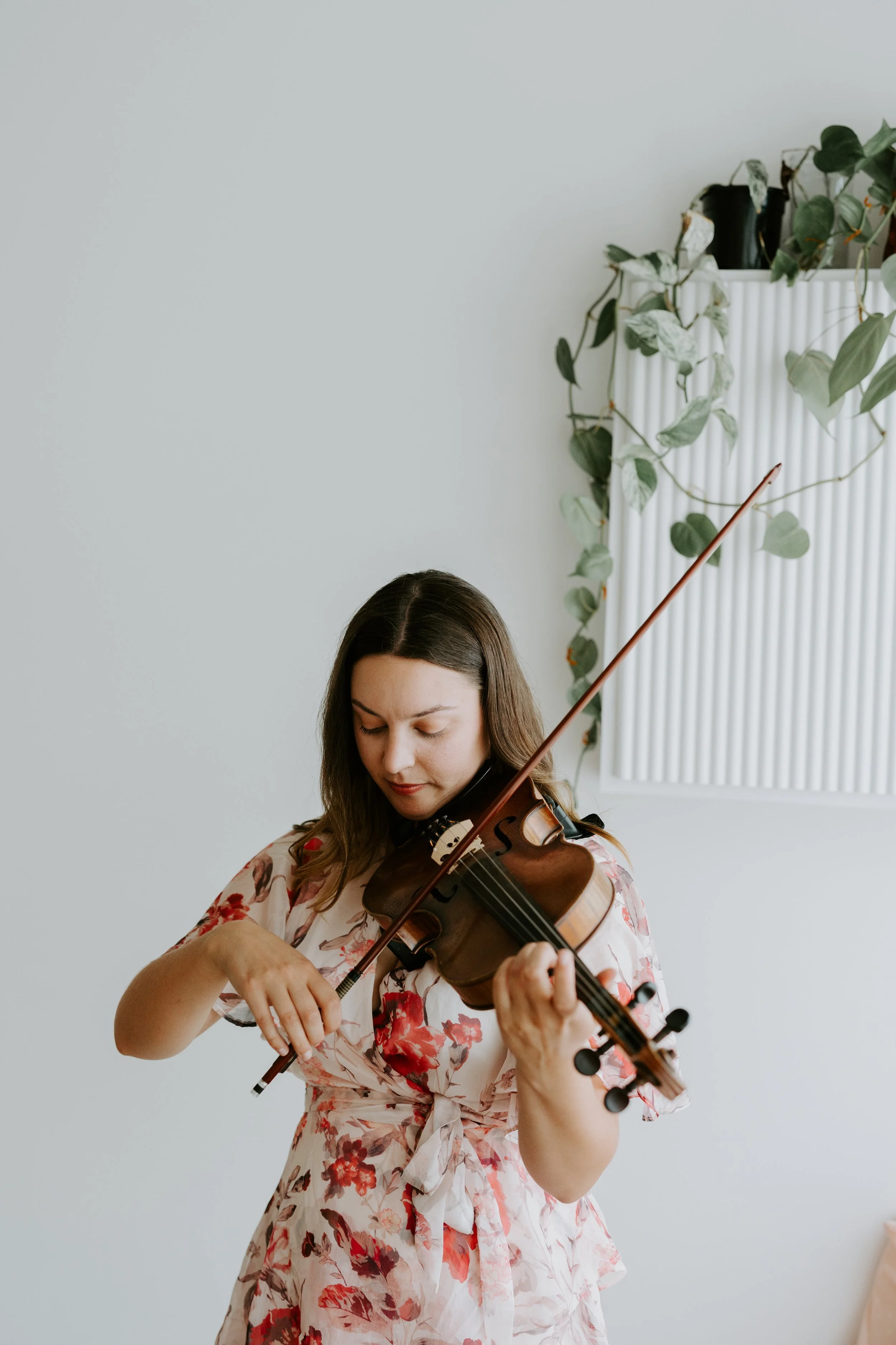 A woman playing a violin indoors with a white wall and green plant in a white ribbed planter behind her.