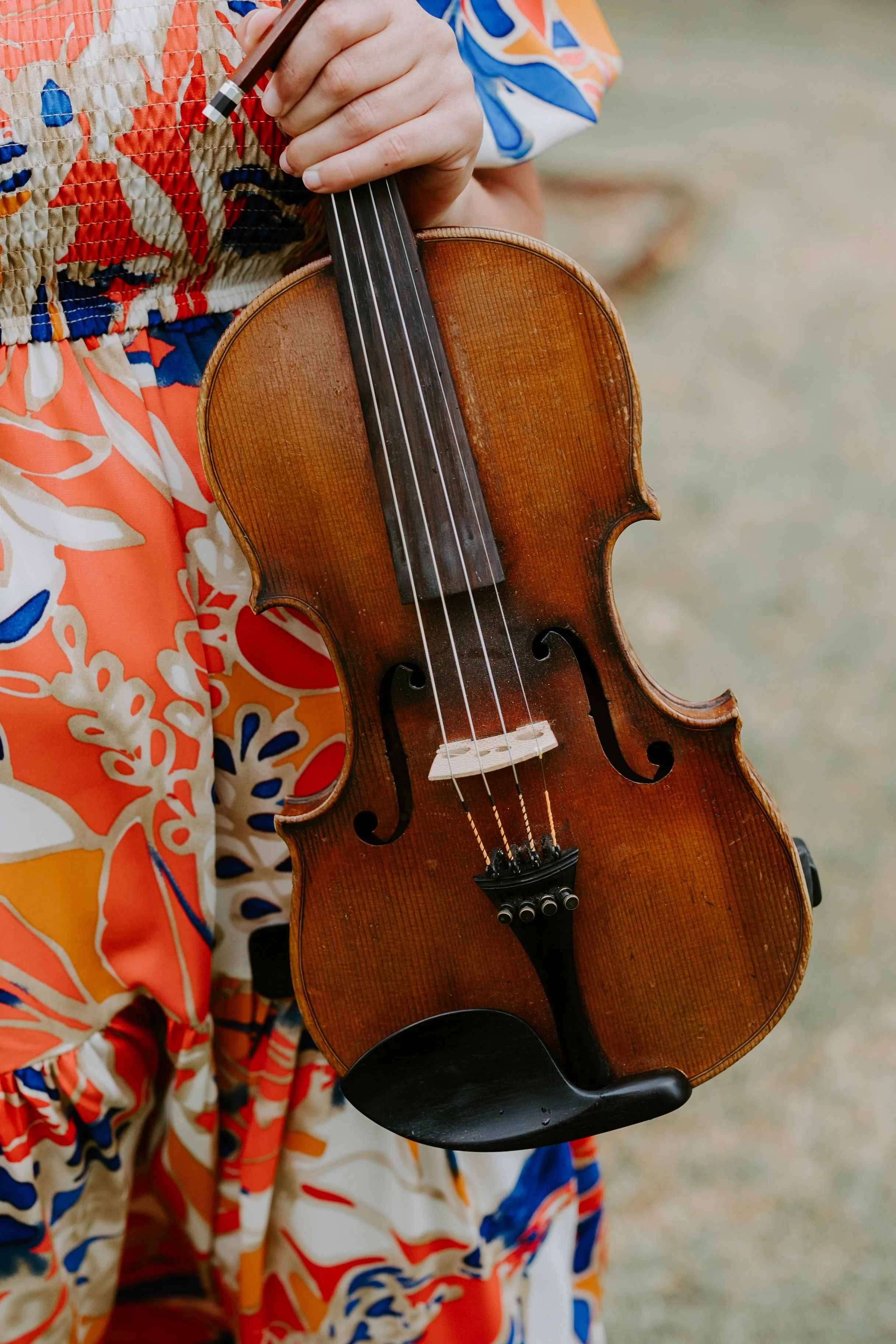 A person wearing a colorful, patterned shirt and pants holding a small, old wooden violin outdoors.