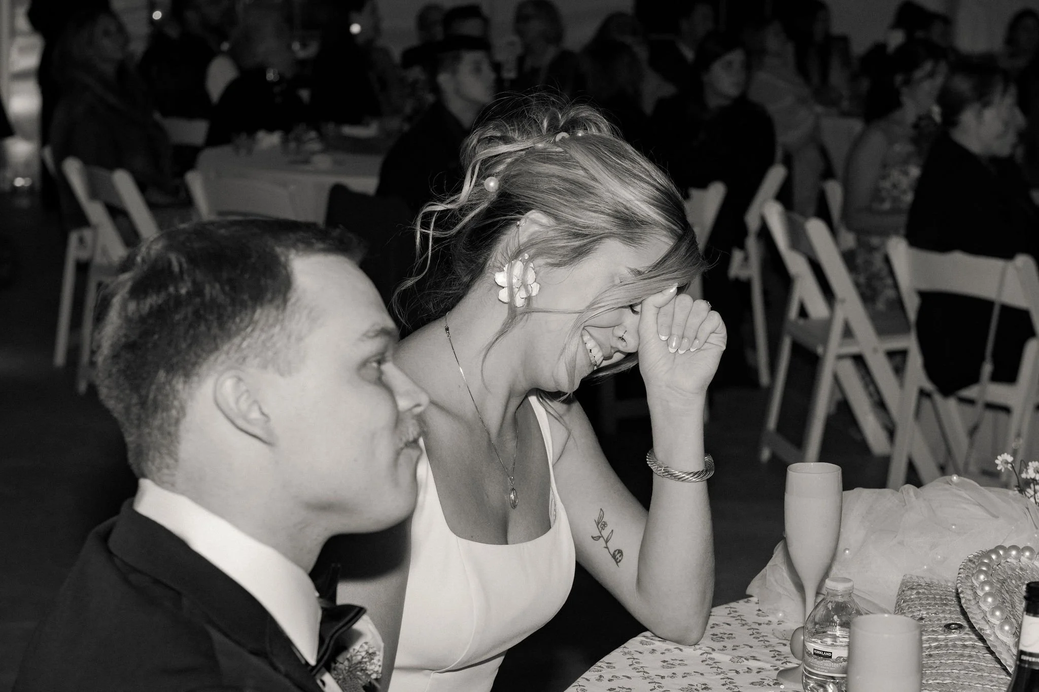 A woman with earrings, a necklace, and a tattoo on her arm is laughing with her hand covering her forehead, sitting next to a man in a tuxedo at a table during a celebration or wedding reception.