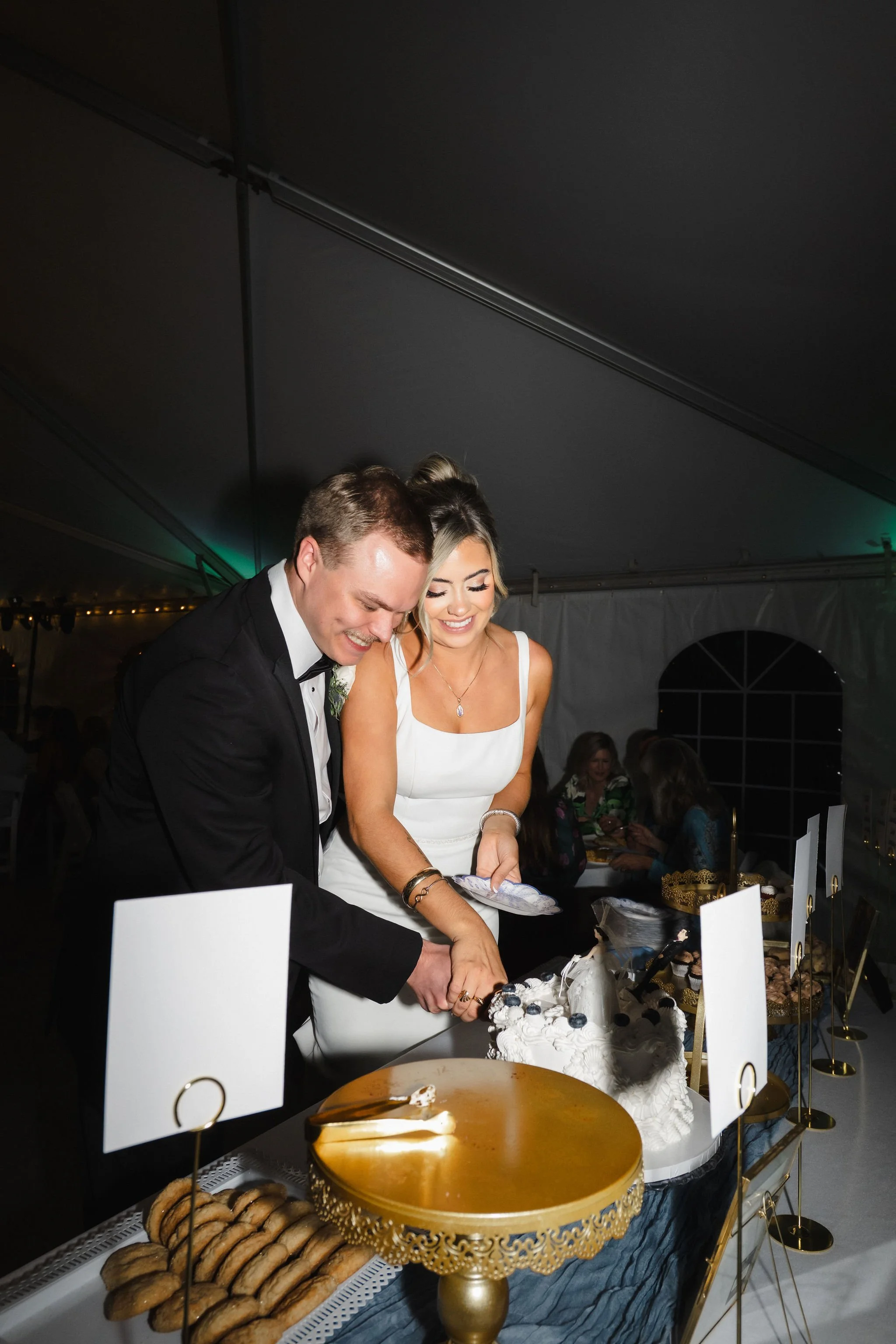 A happy bride and groom cutting their wedding cake together at a reception tent with a dessert table in the background.