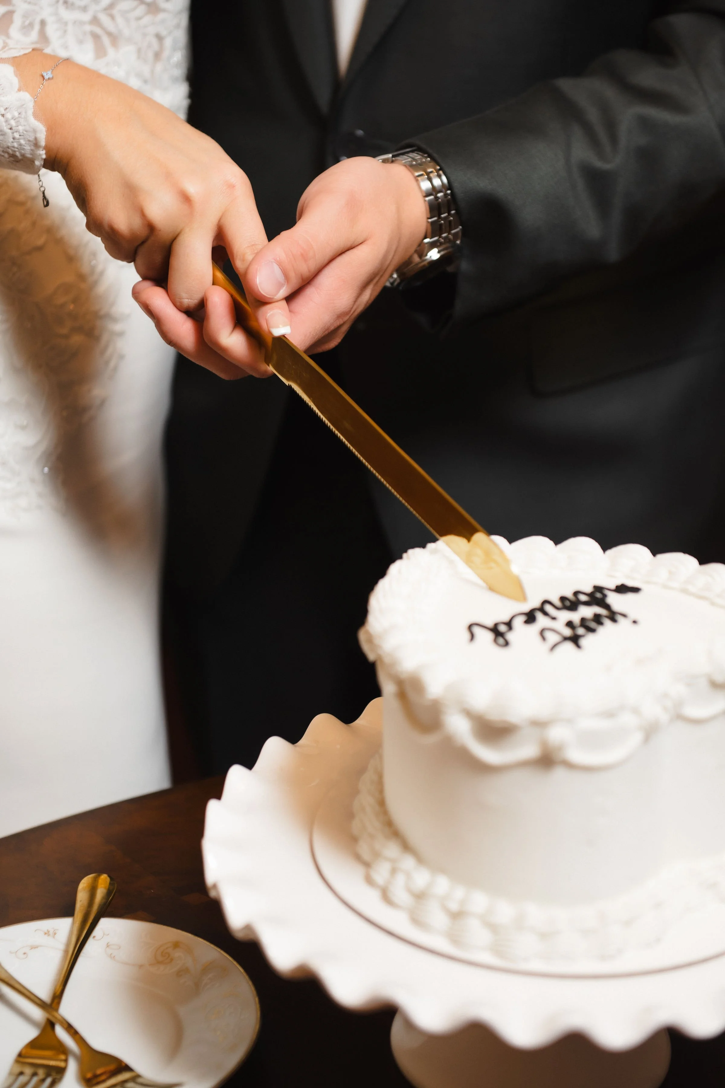 A couple cuts a wedding cake together, with the bride's hand holding the knife and the groom's hand guiding it, on a silver knife with a gold blade, in front of a white wedding cake.