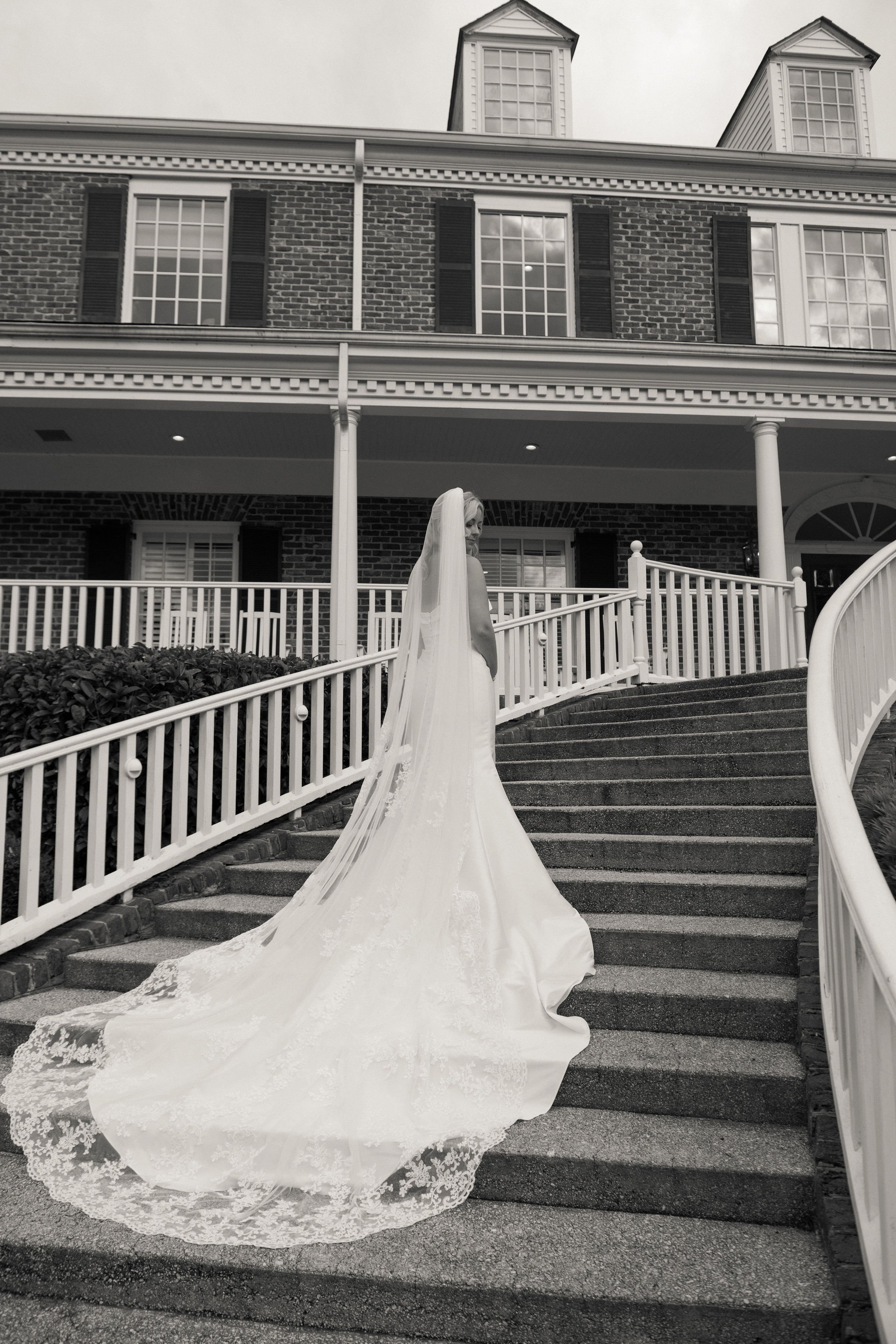 A bride in a wedding gown and veil standing on the steps of a large brick house with a white railing, looking over her shoulder.