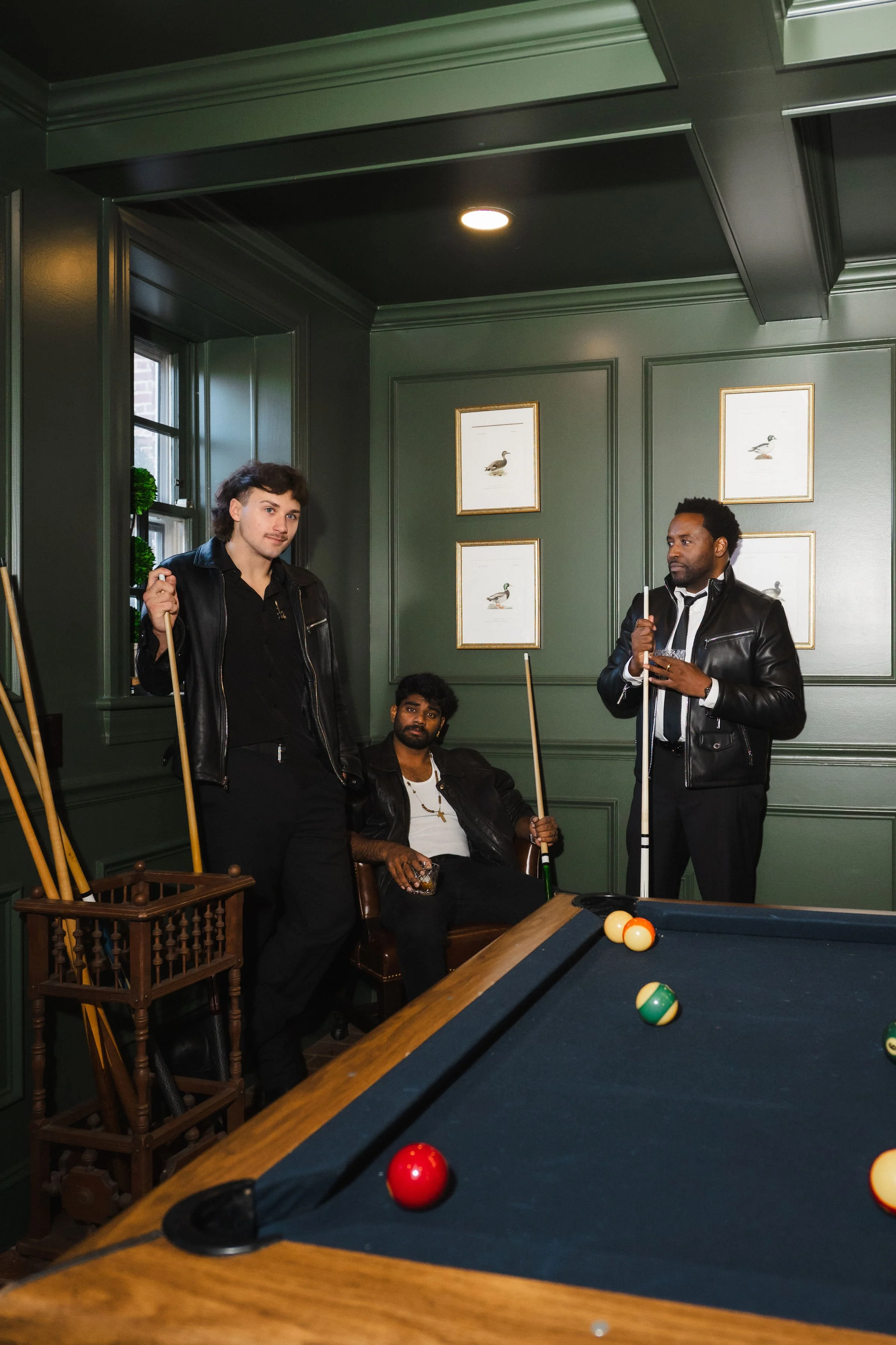 Three men in leather jackets playing pool in a dimly lit green-walled room with framed bird pictures.