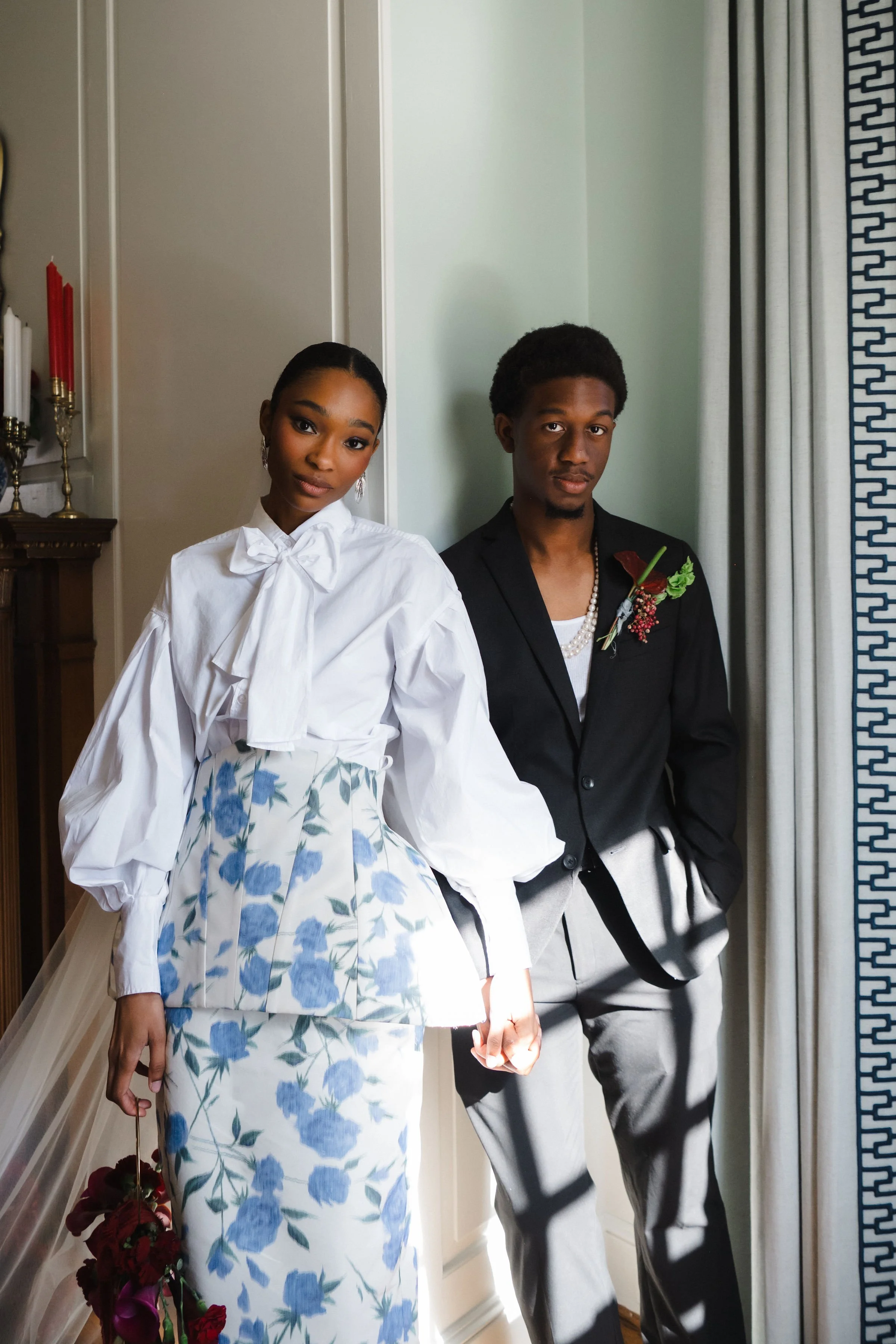 A young woman and man dressed in formal attire standing inside a room near a window with patterned curtains. The woman is wearing a white blouse with a bow tie and a floral skirt, holding a bouquet of purple and red flowers. The man is wearing a black jacket with a boutonniere, pearl necklaces, and grey pants, with his hands in his pockets.