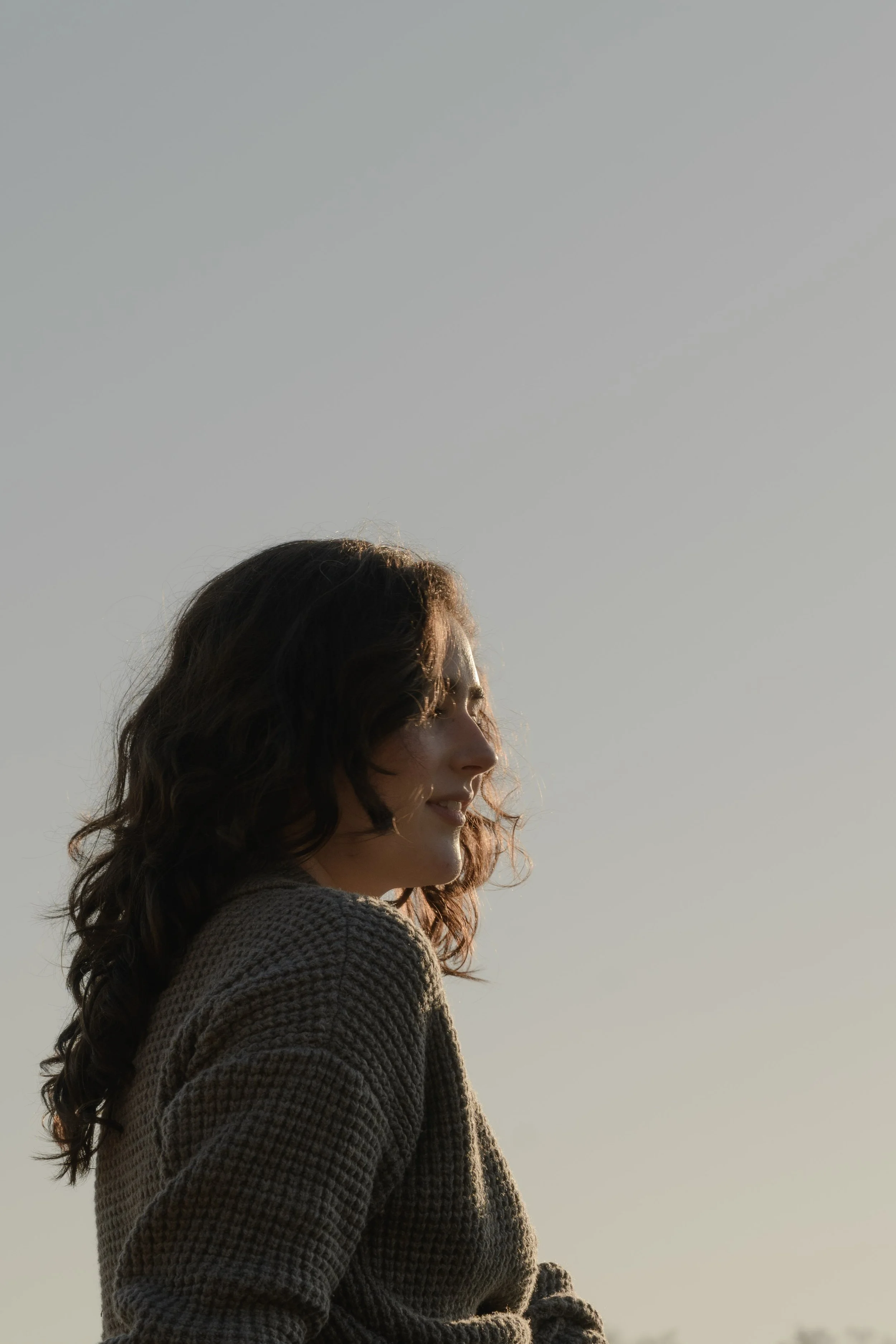 Side profile of a smiling woman with wavy brown hair, wearing a knitted sweater, outdoors during natural daylight.