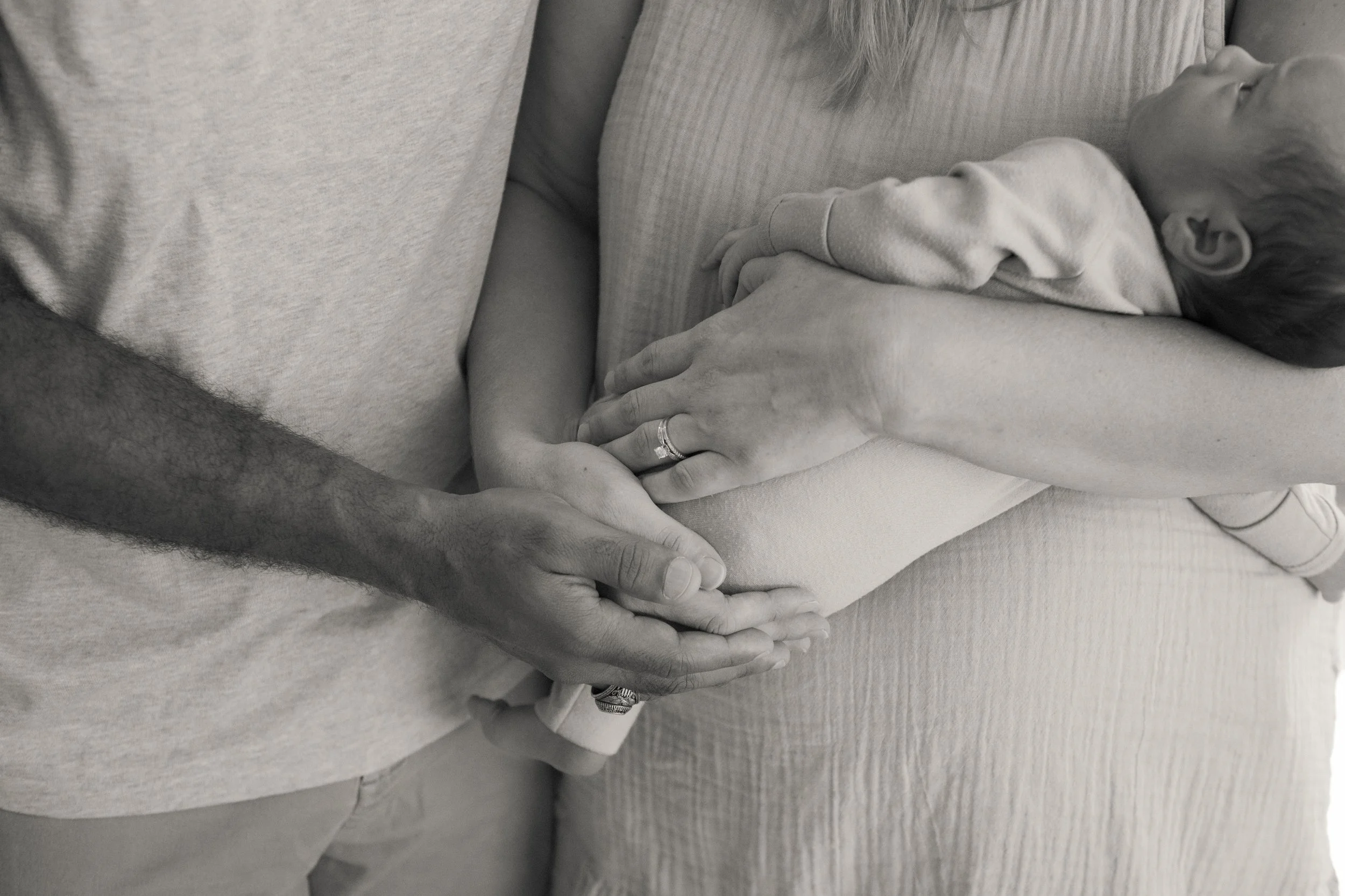 A black and white photo showing a man and woman holding a baby's hand, with the baby lying on the woman. The baby is nude from the waist up, resting on the woman's lap, with a blanket covering the lower part of the baby. The man has a wedding ring on