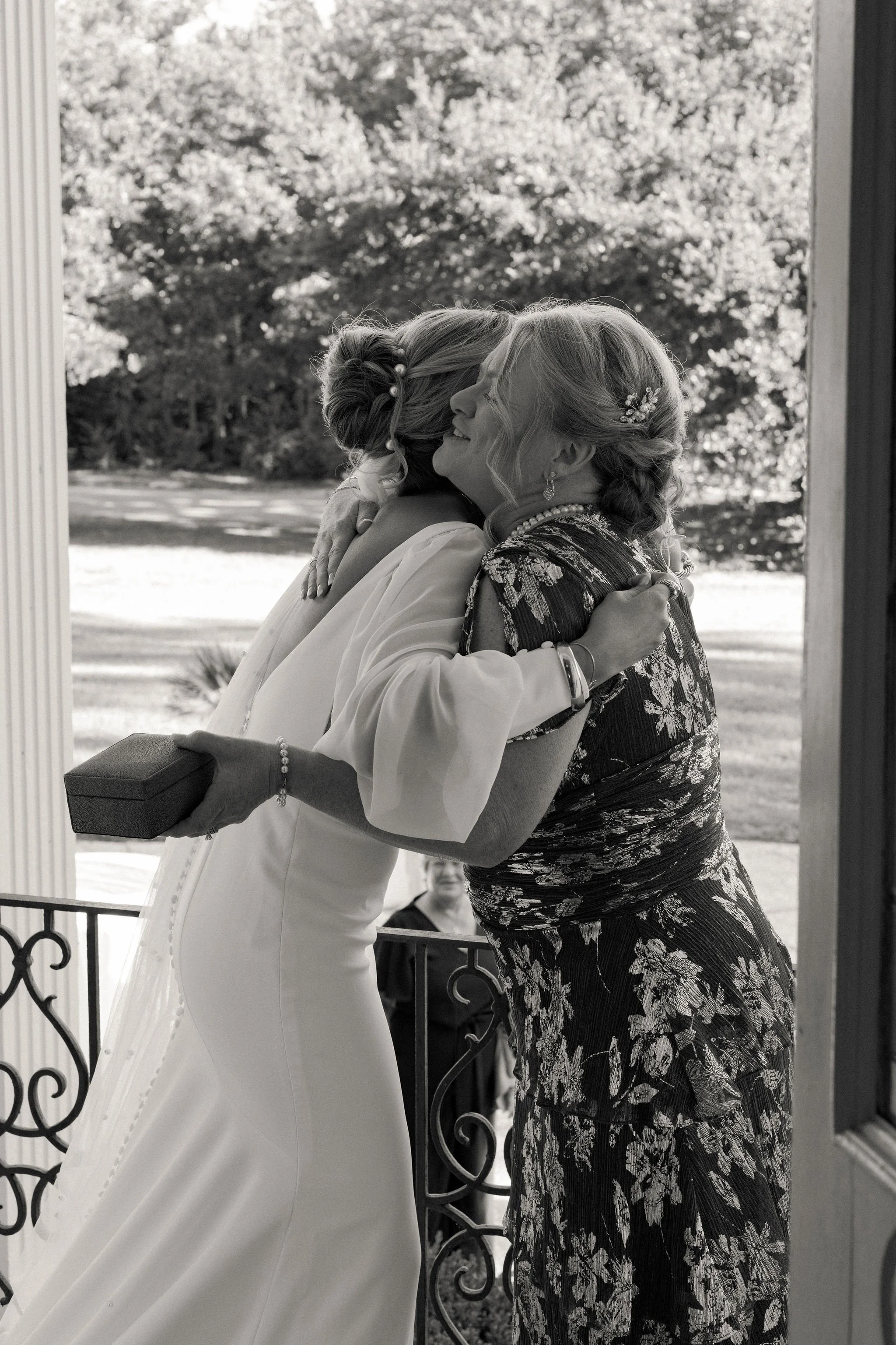 A bride hugging an older woman, possibly her mother, on a porch during a wedding. The bride is holding a small box, and both are smiling warmly.