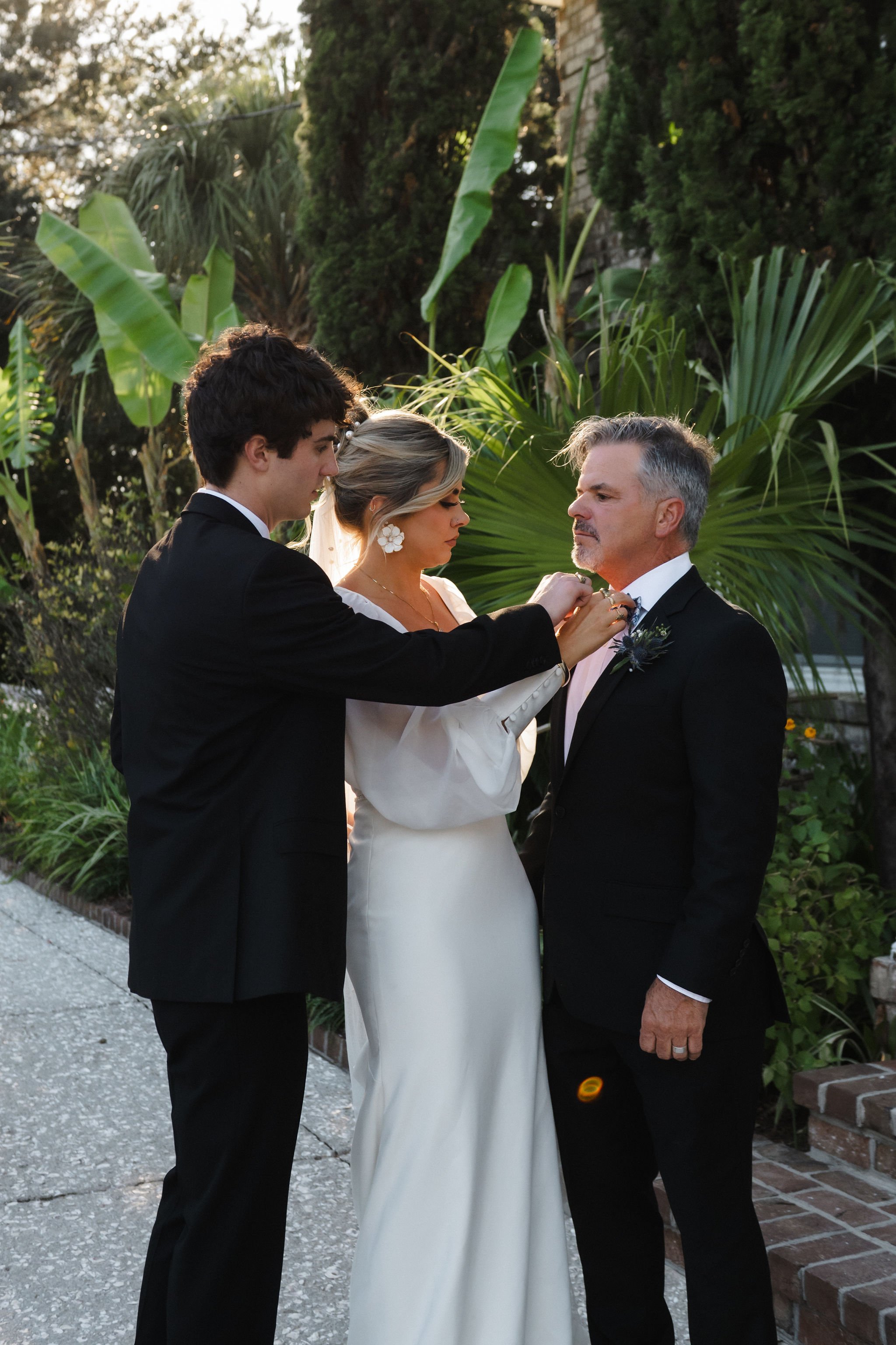 A woman in a white dress with flower earrings stands between two men in black suits outdoors, as one man adjusts his tie and the woman looks down, surrounded by greenery.