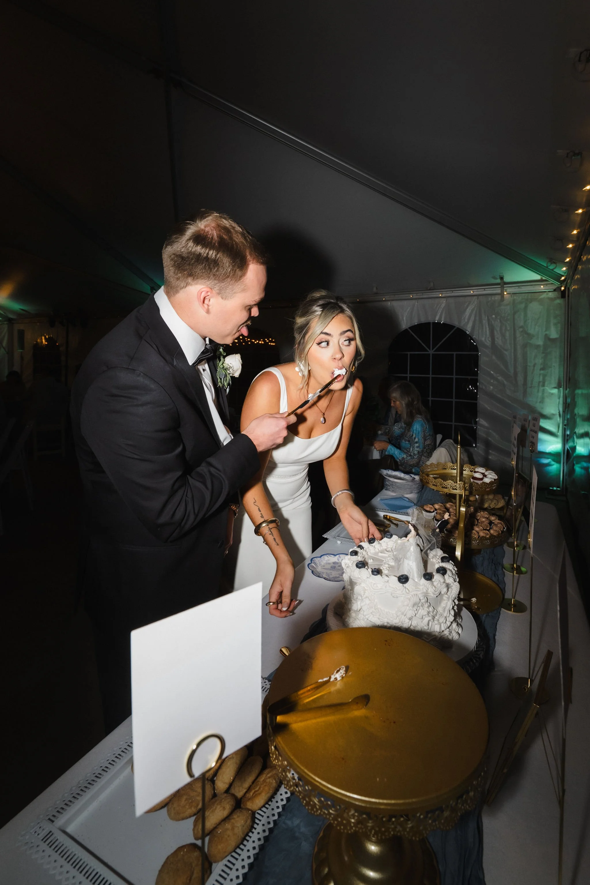 Bride and groom feeding each other cake at a wedding reception under a large tent.