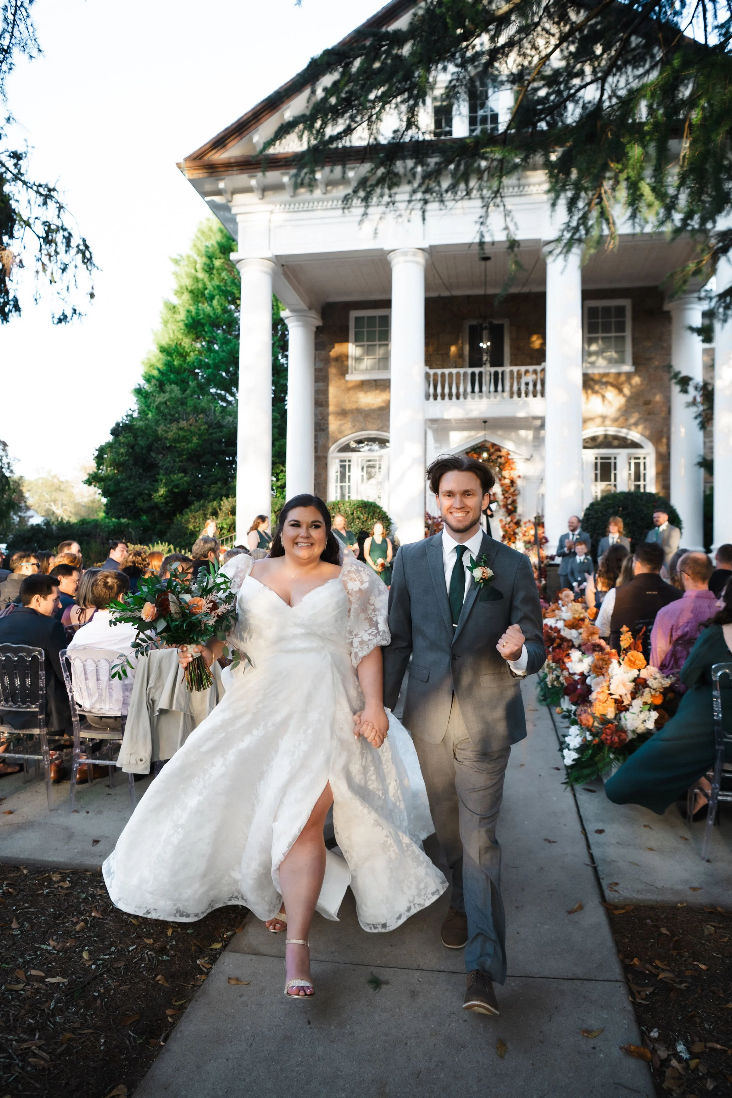 A bride and groom walk hand in hand down the aisle of an outdoor wedding reception, smiling happily. The bride is in a white wedding dress holding a bouquet, and the groom is in a gray suit with a tie. The reception area is decorated with colorful fl