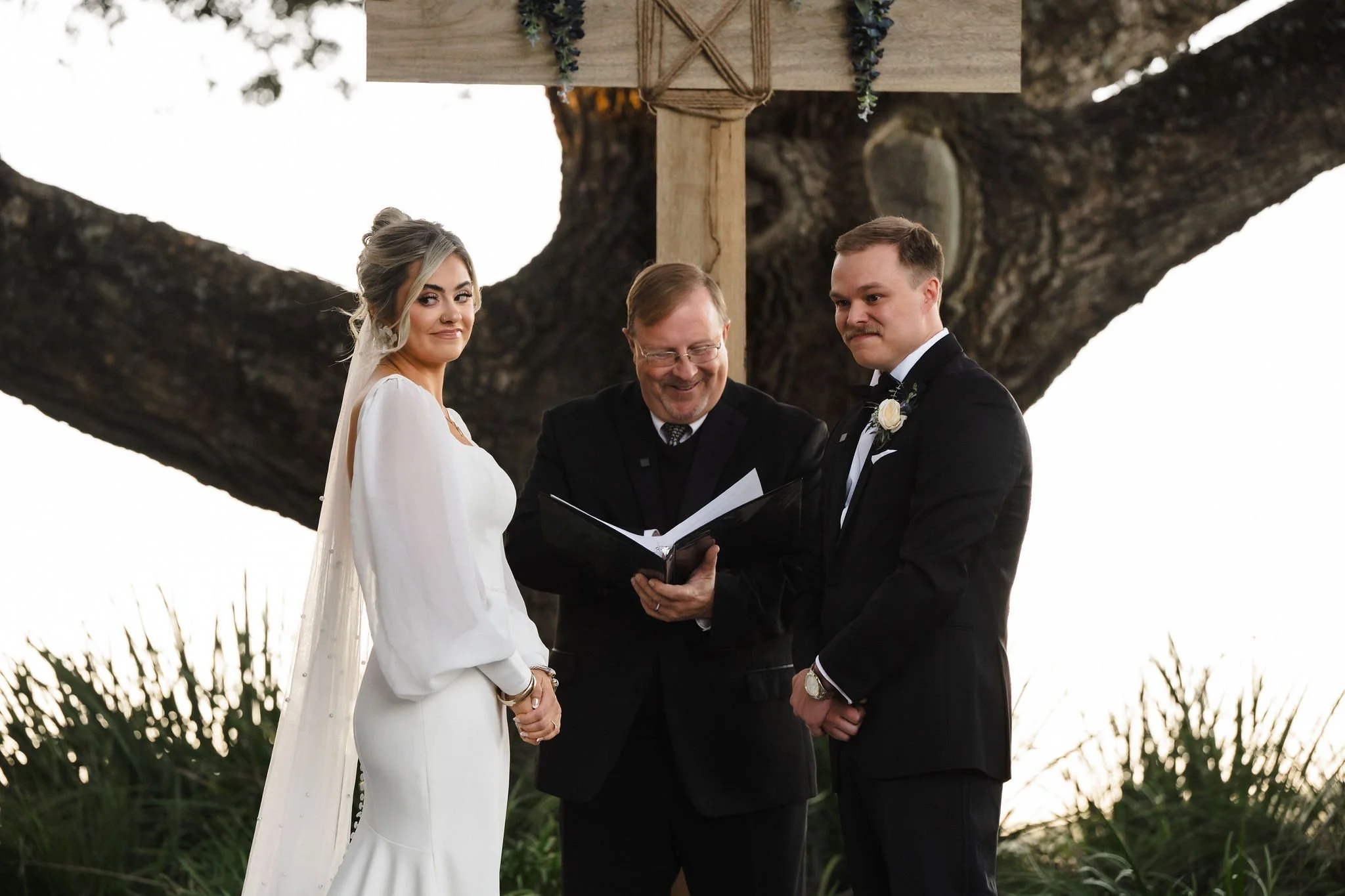 A bride and groom stand outdoors under a large tree during their wedding ceremony, with a male officiant reading from a book between them.