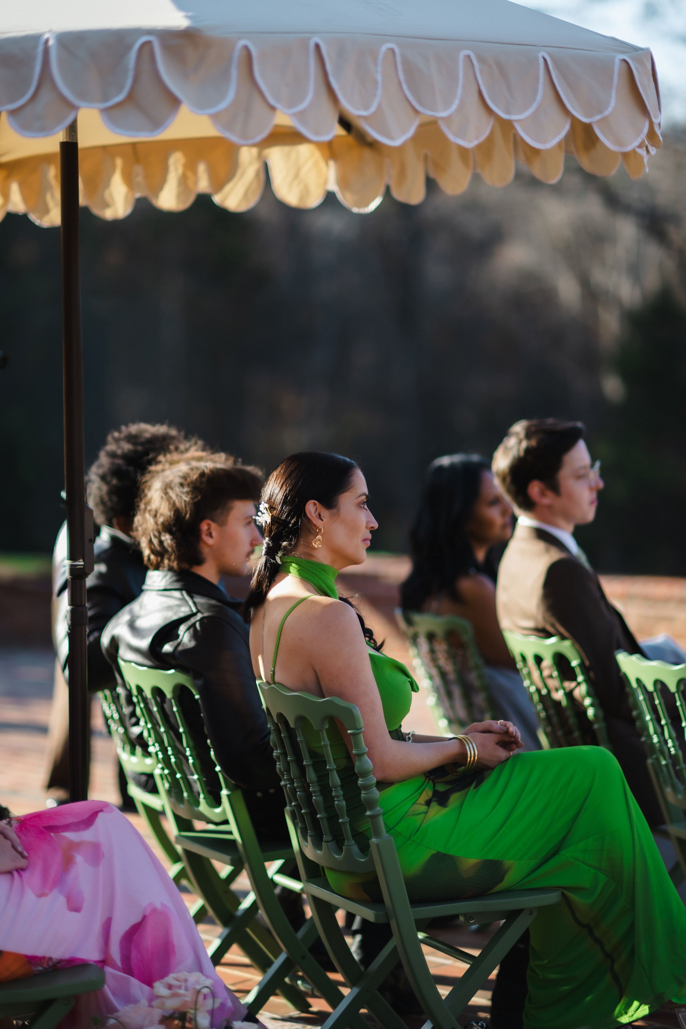 People sitting under a cream-colored umbrella at an outdoor event, with some wearing formal and colorful attire.
