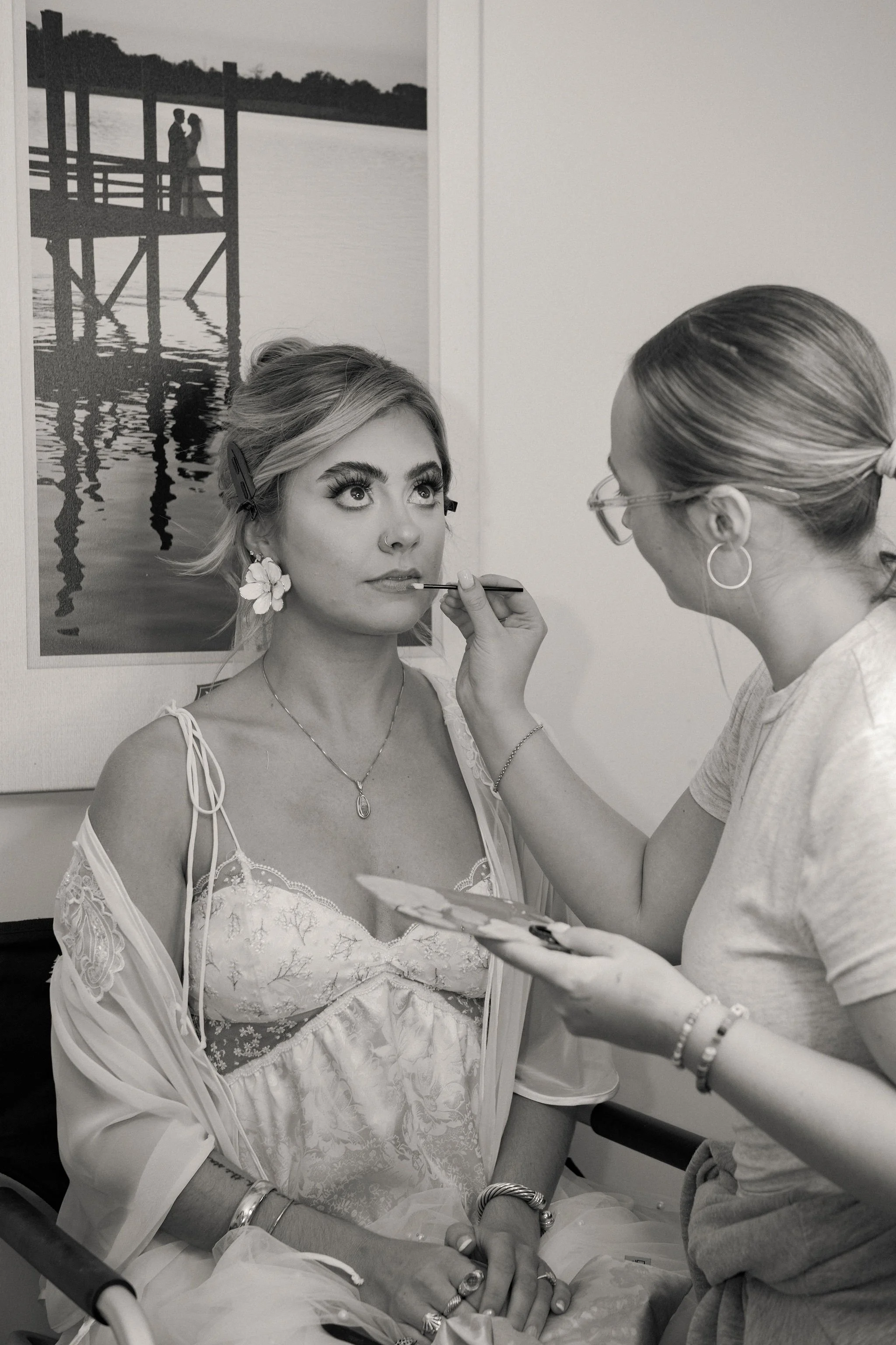 A woman with light-colored hair styled up, sitting in a chair while another woman applies makeup on her lips. The woman getting makeup has floral earrings, a necklace, and wears a light-colored dress with lace details, along with several bracelets an