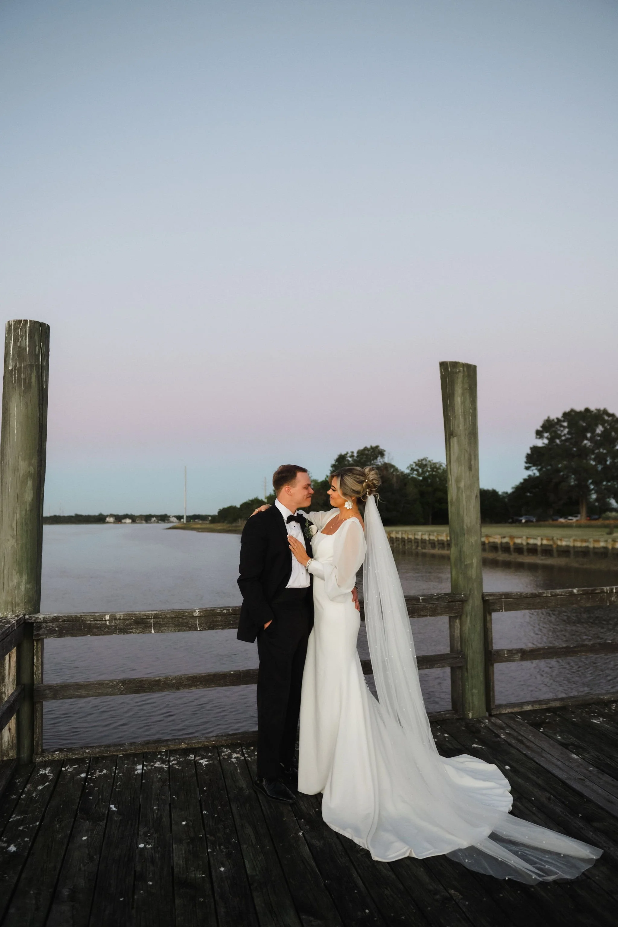 A bride and groom in wedding attire on a wooden dock by the water, embracing at sunset with a pastel sky in the background.