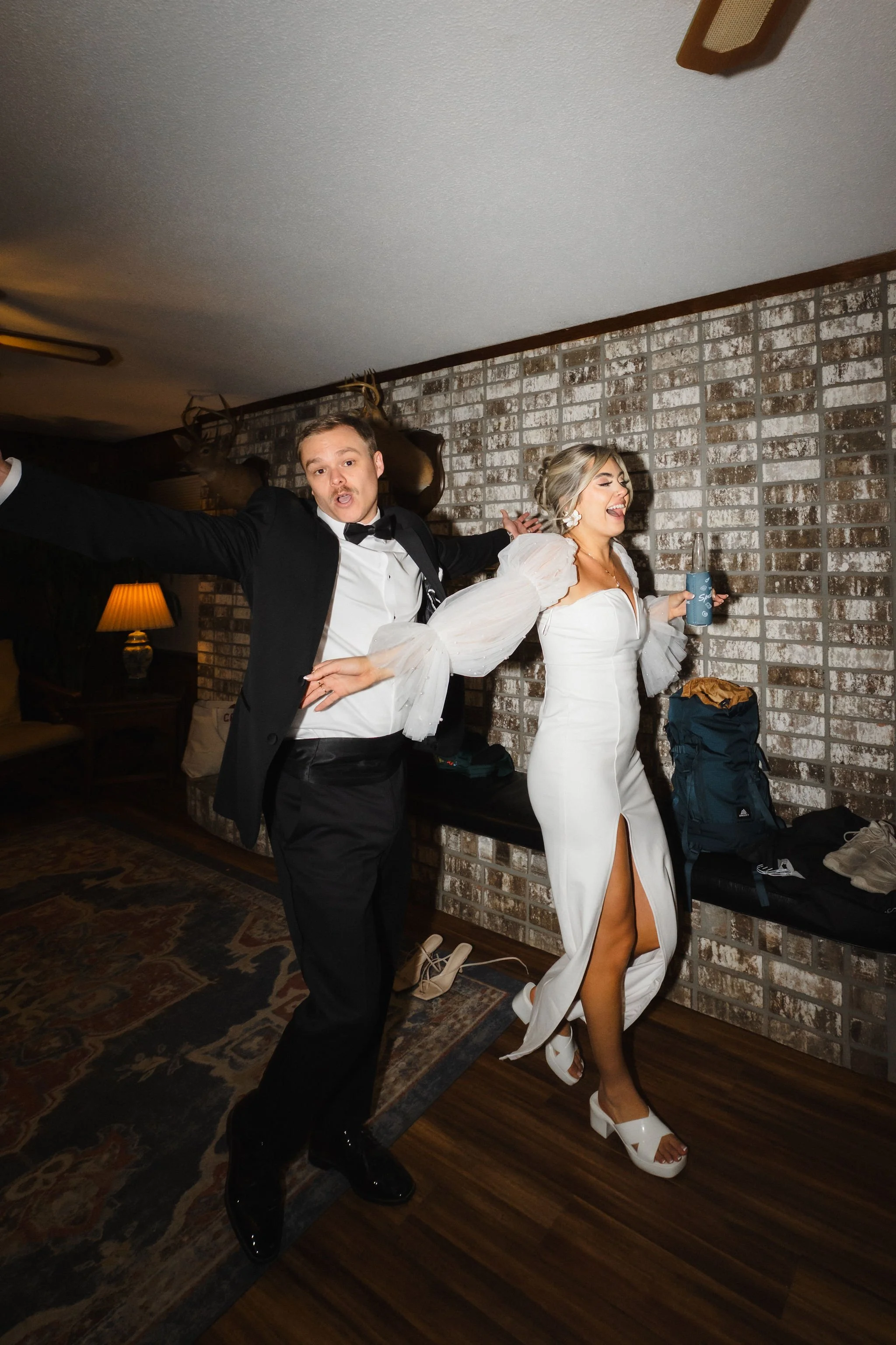 A man and woman dance happily at a party. The man is dressed in a tuxedo and the woman in a white dress with a high slit, both enjoying themselves.