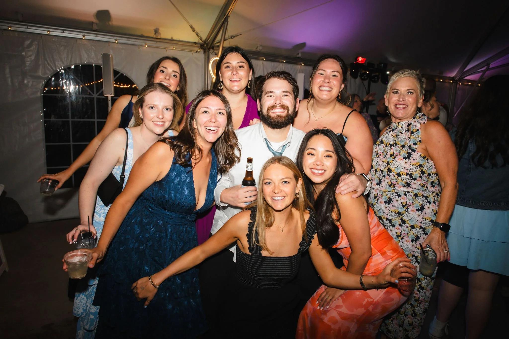 Group of women and one man at a party, smiling and holding drinks, inside a tent with string lights.