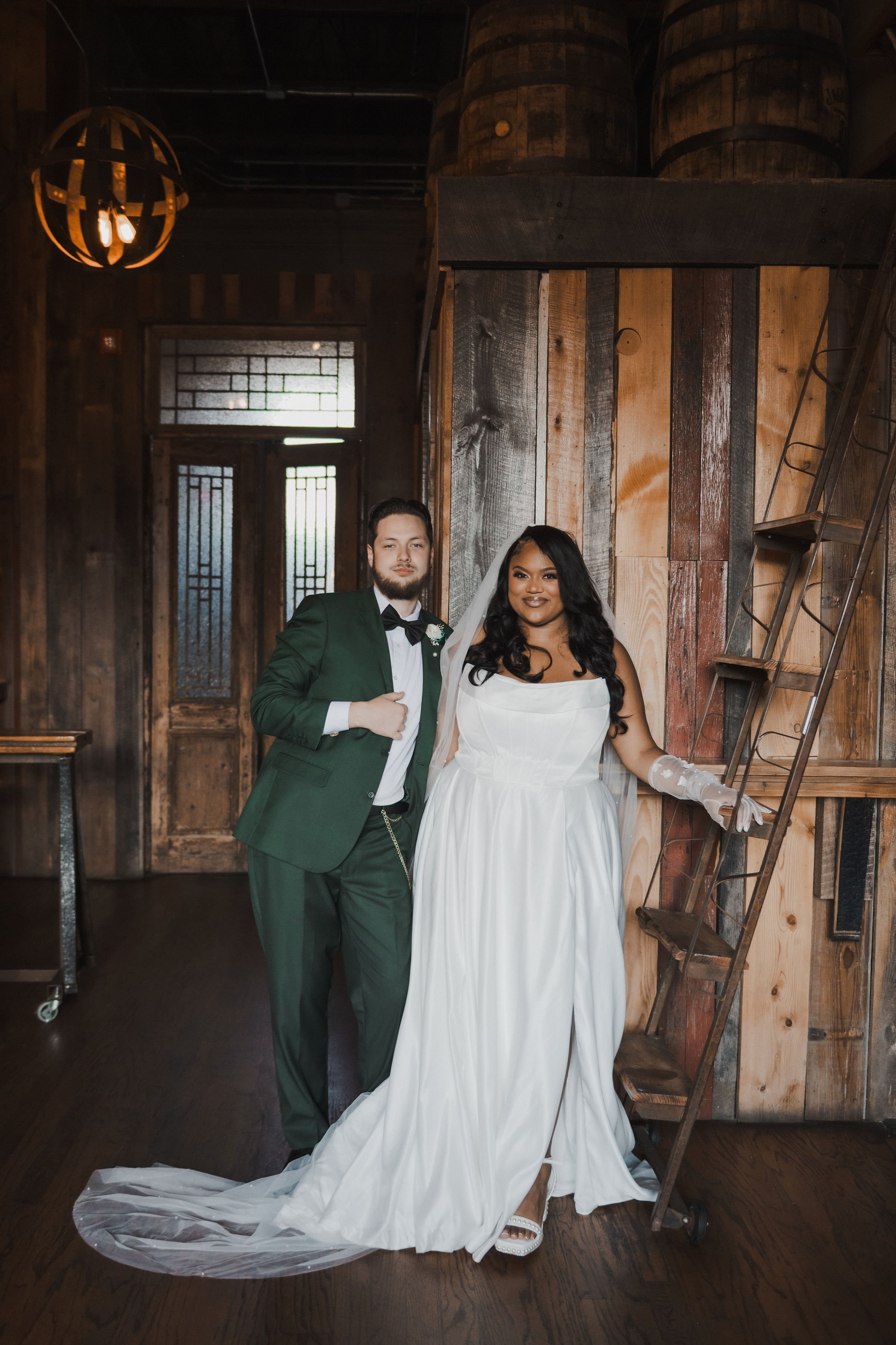 A newlywed couple in wedding attire standing indoors with rustic wooden walls and stairs.