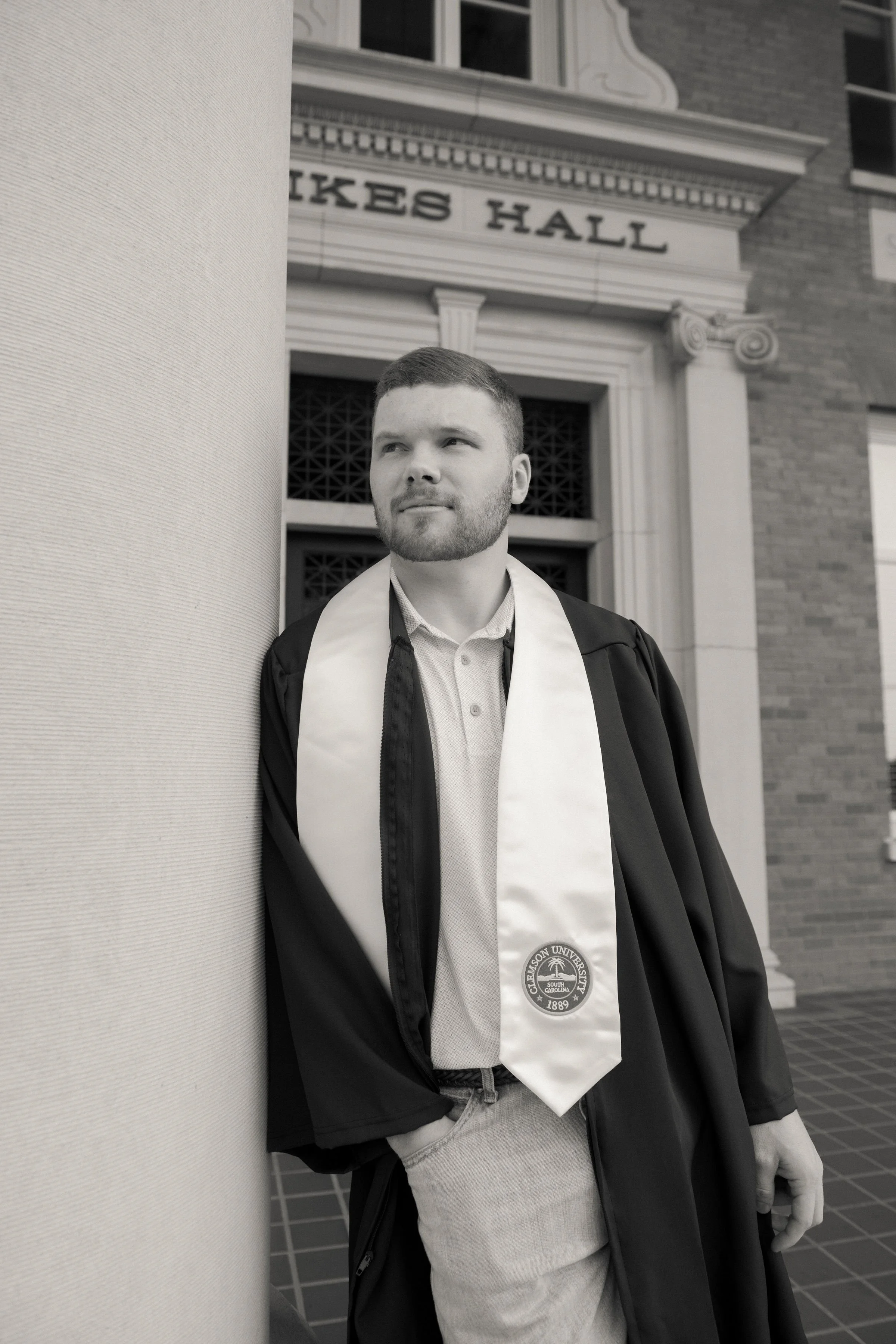A young man in a graduation gown and sash standing outdoors near a column in front of a building labeled 'HICKES HALL'.