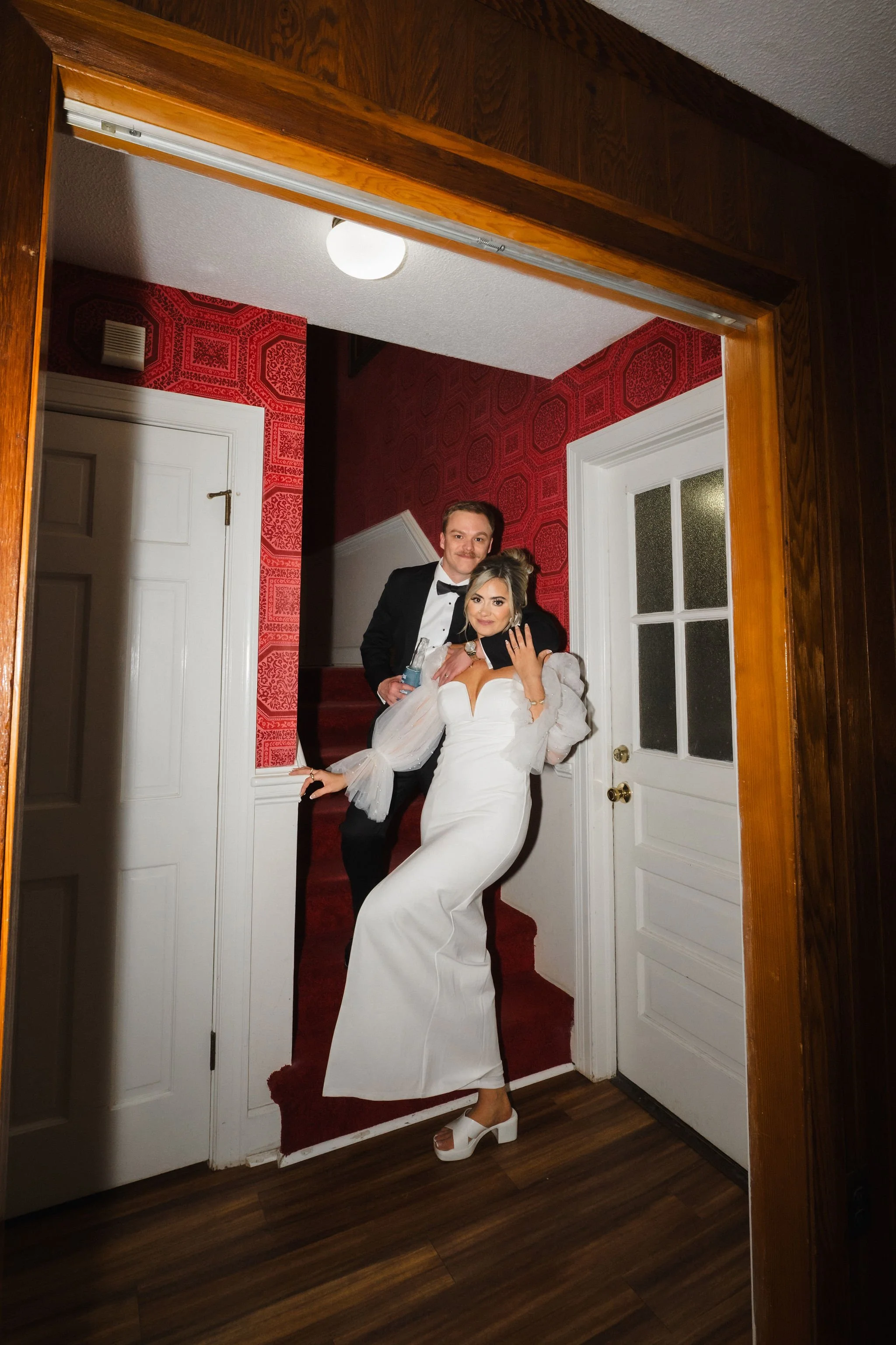 A couple in formal attire posing in a small hallway with red patterned wallpaper. The woman is in a white dress and white high-heeled shoes, while the man is in a black tuxedo. They are standing at the bottom of a staircase, smiling at the camera.