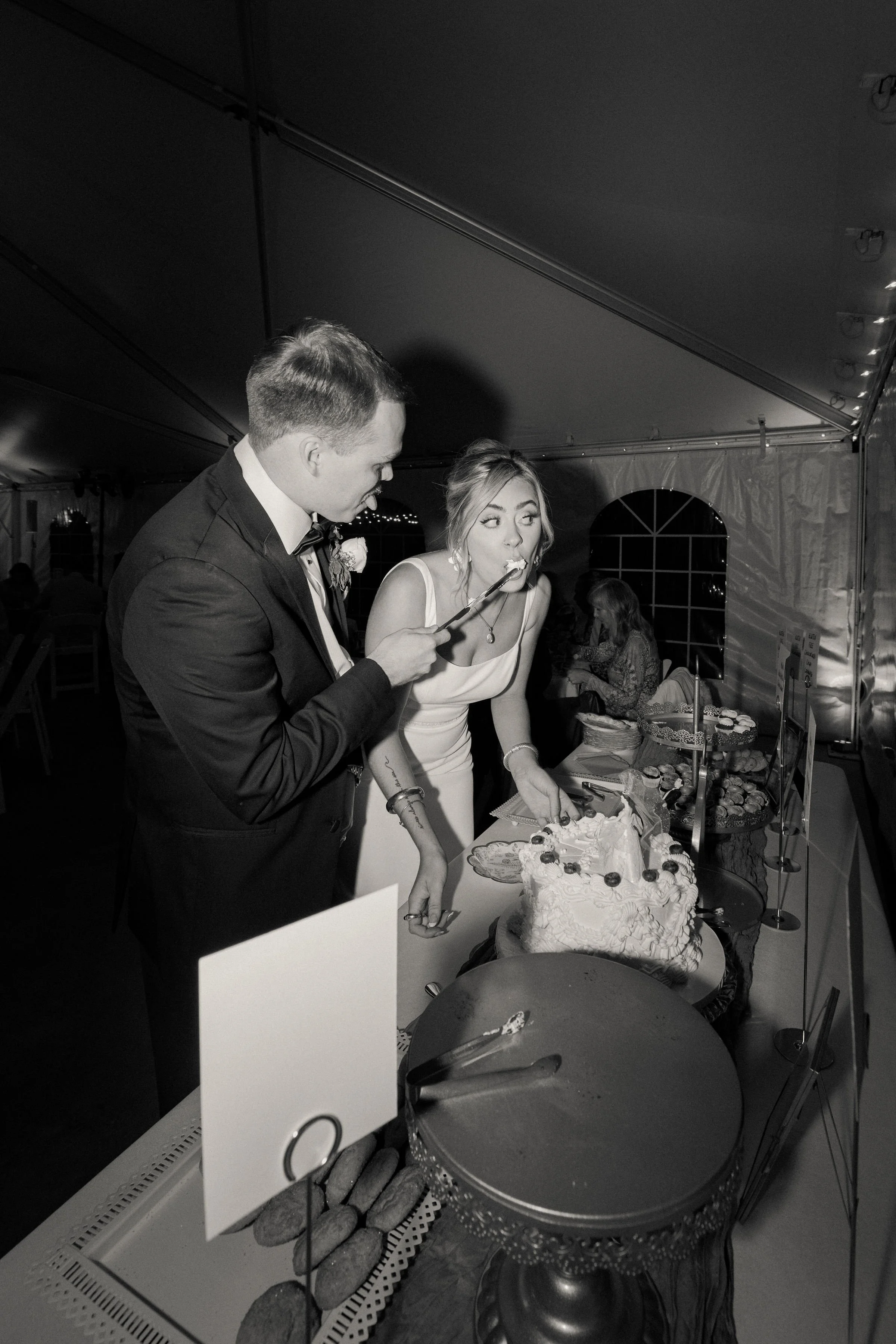 Black and white photo of a bride and groom cutting their wedding cake at a reception, with the groom using a cake server and the bride holding a piece of cake on a fork