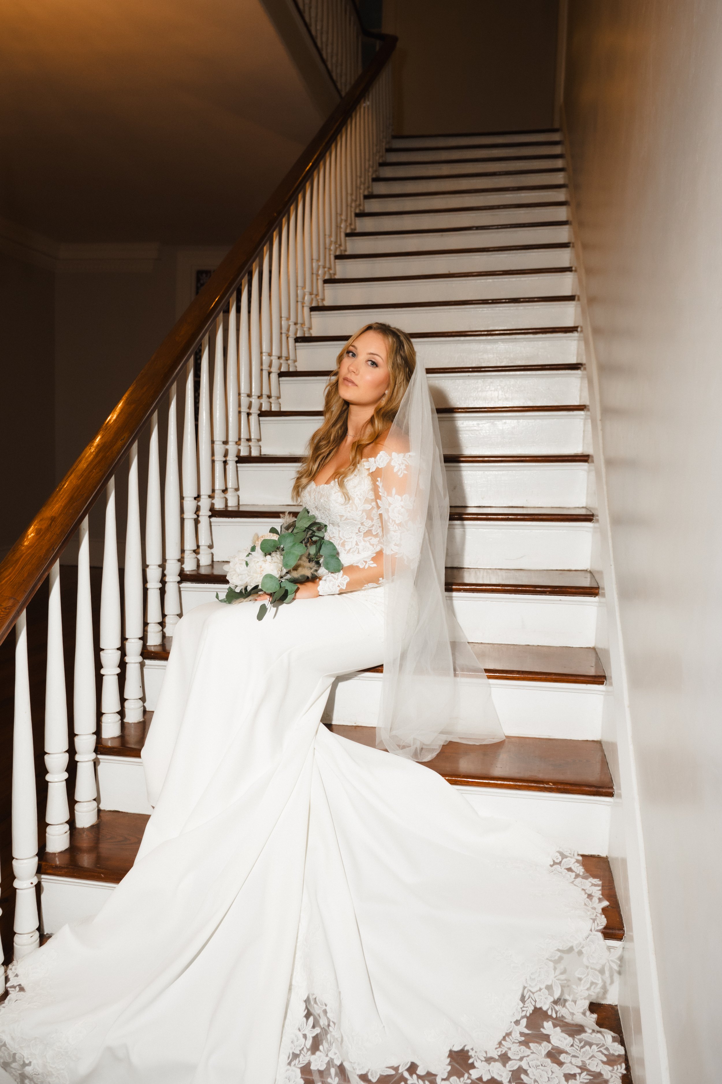 A bride in a white wedding dress with lace sleeves and a long train, sitting on the stairs with a bouquet of white flowers and greenery, wearing a veil, looking at the camera.