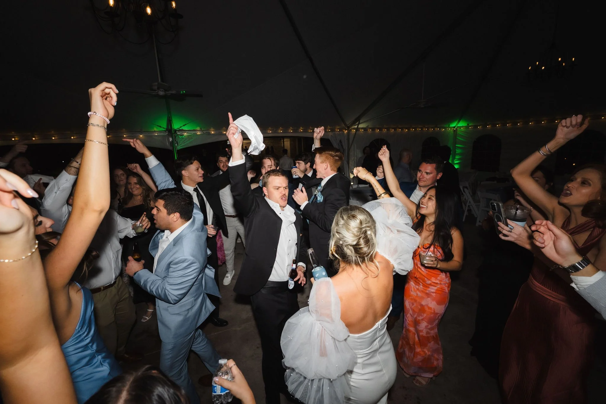 People dancing and celebrating at a wedding reception inside a tent, with some holding drinks and raising their hands in the air.