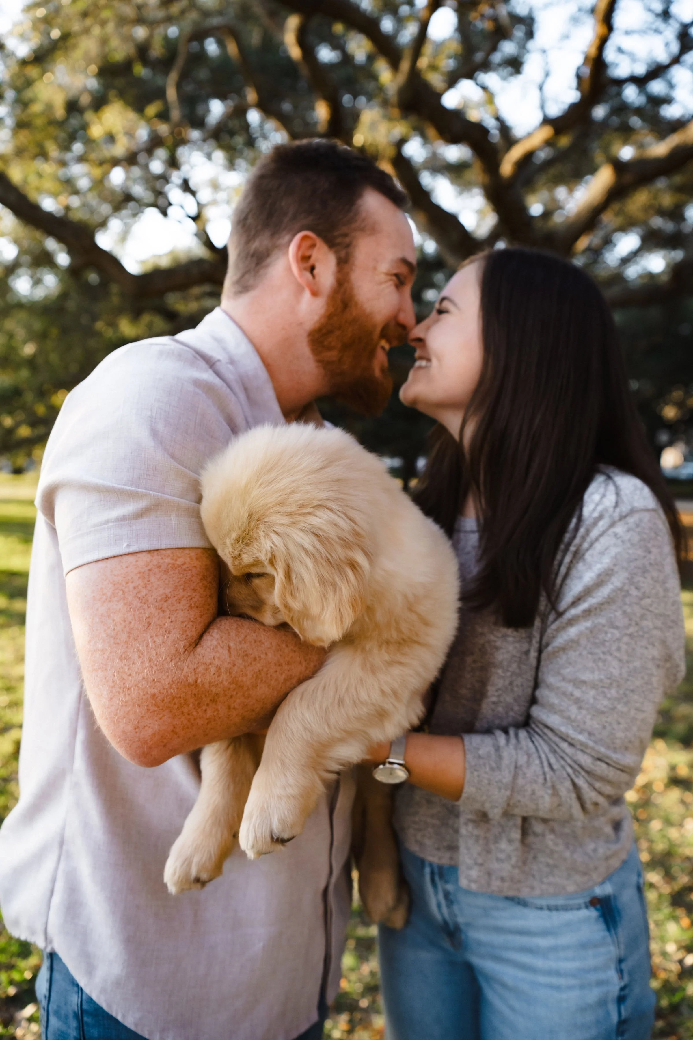 A couple smiling and touching noses while holding a golden retriever puppy in a park during daytime.