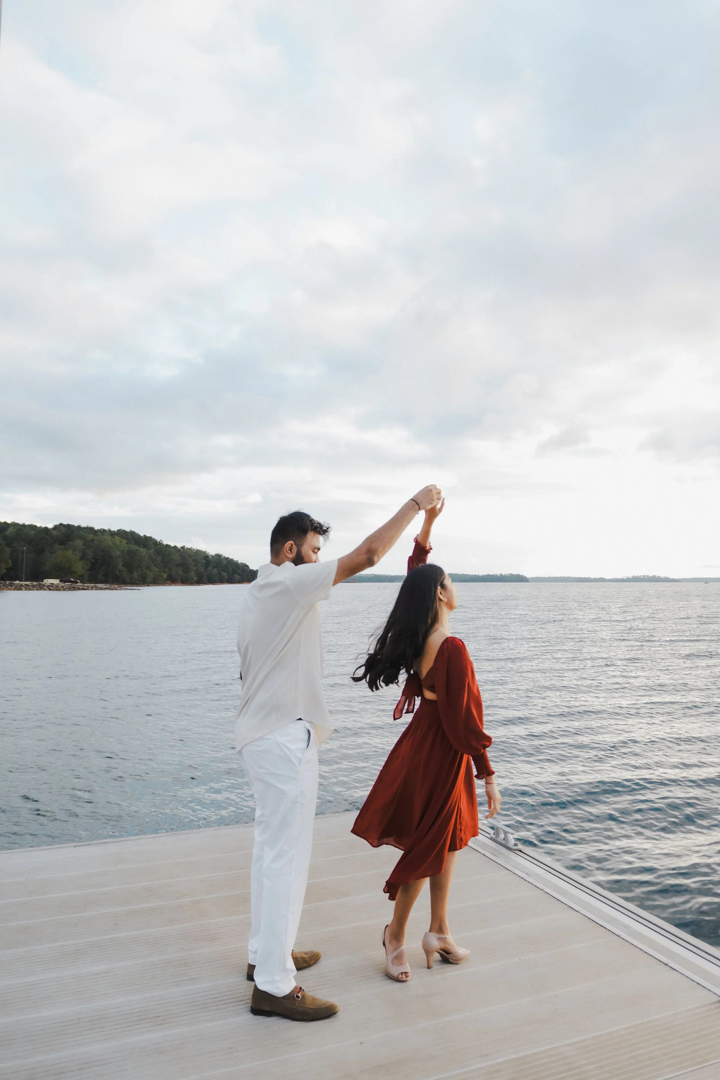 A couple dancing on a dock by the water during sunset, with a cloudy sky and distant shoreline in the background.