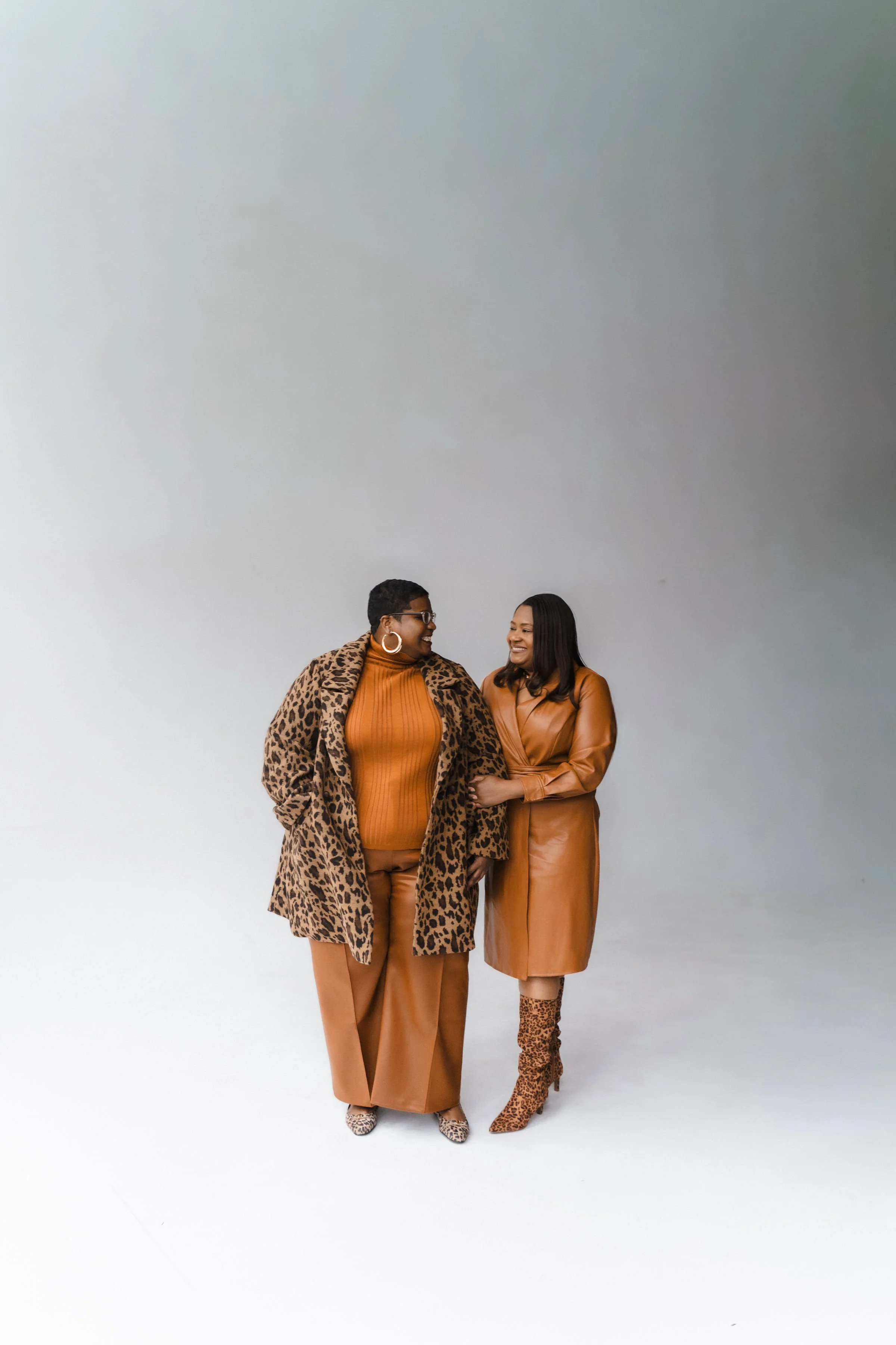 Two women dressed in brown and animal print outfits standing close together and smiling against a neutral background.