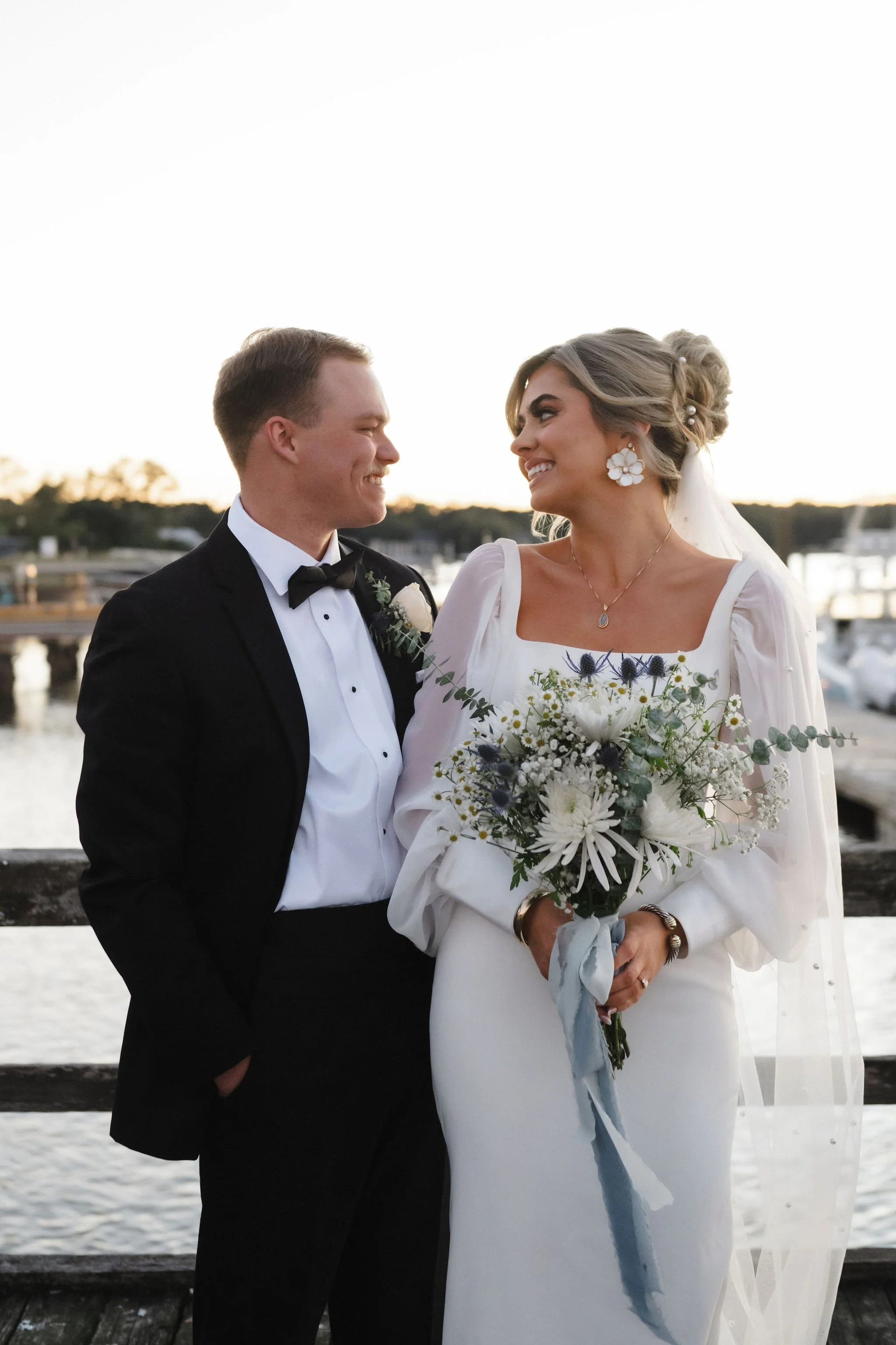 Bride and groom smiling at each other on a dock by the water during sunset, with boats and trees in the background.