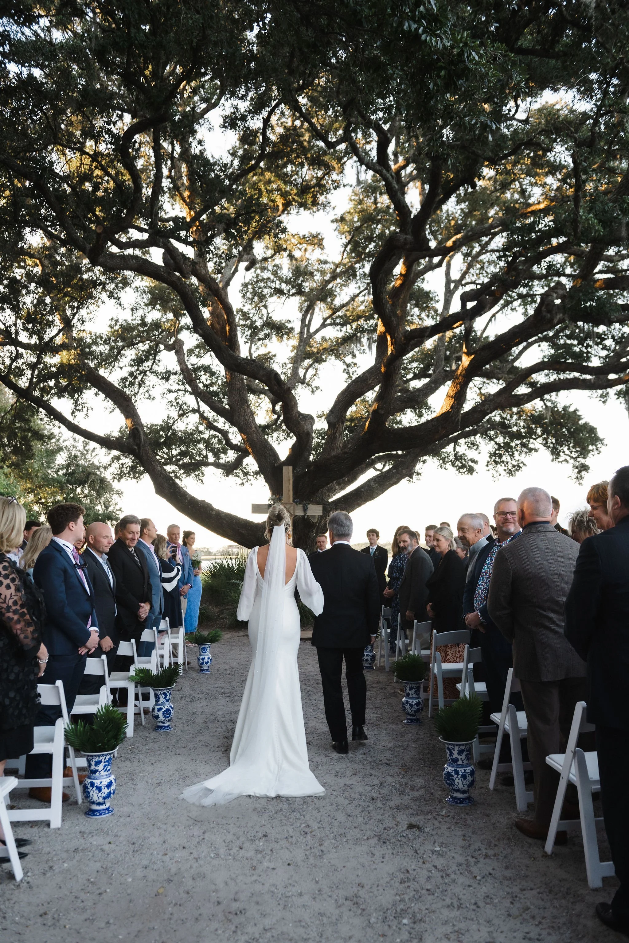 A wedding ceremony outdoors under a large tree, with the bride walking down the aisle accompanied by a person, possibly her father, as guests stand on either side.