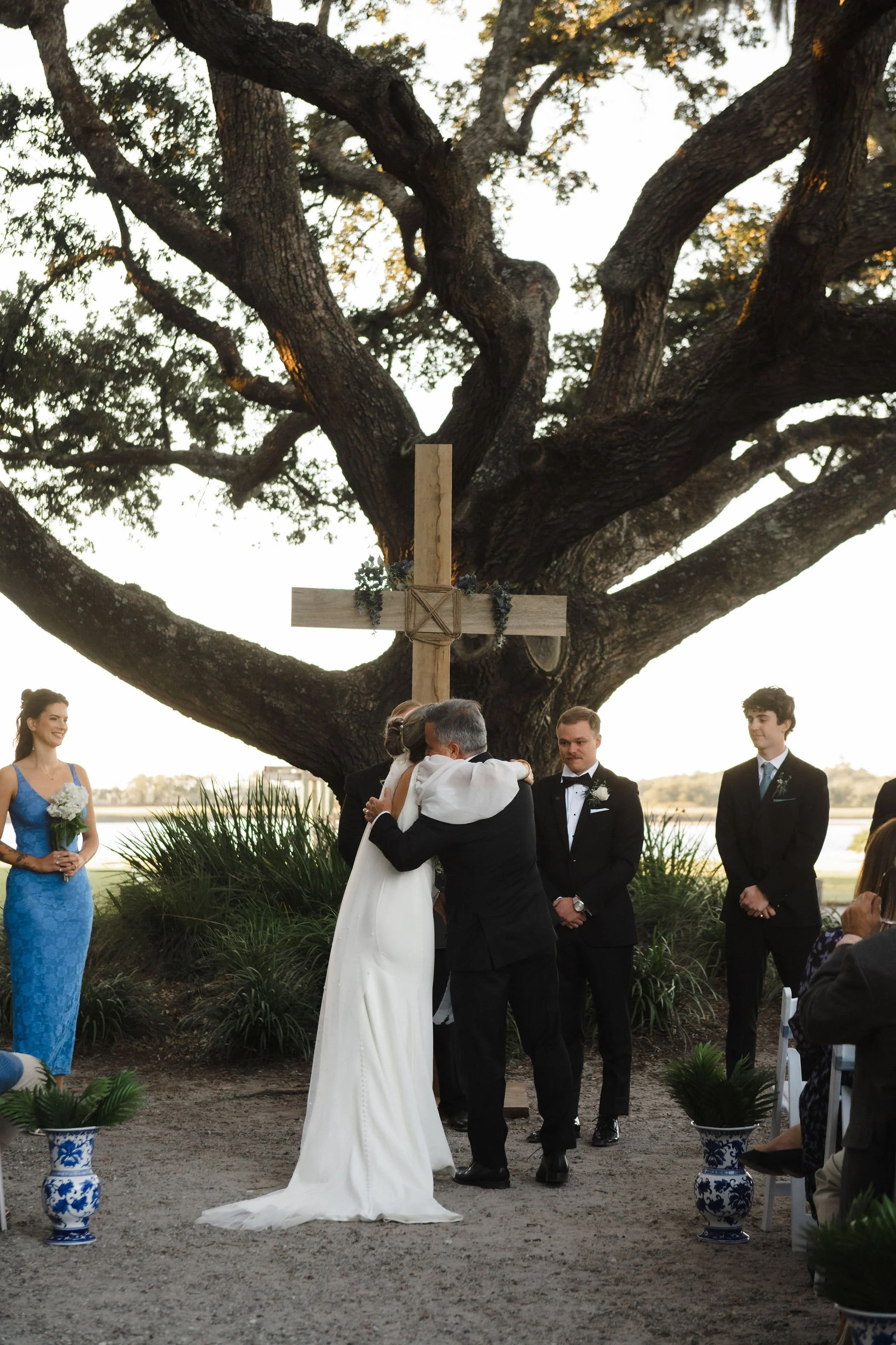 A wedding ceremony outdoors under a large tree with a cross, featuring a bride and groom hugging, surrounded by attendants and guests.