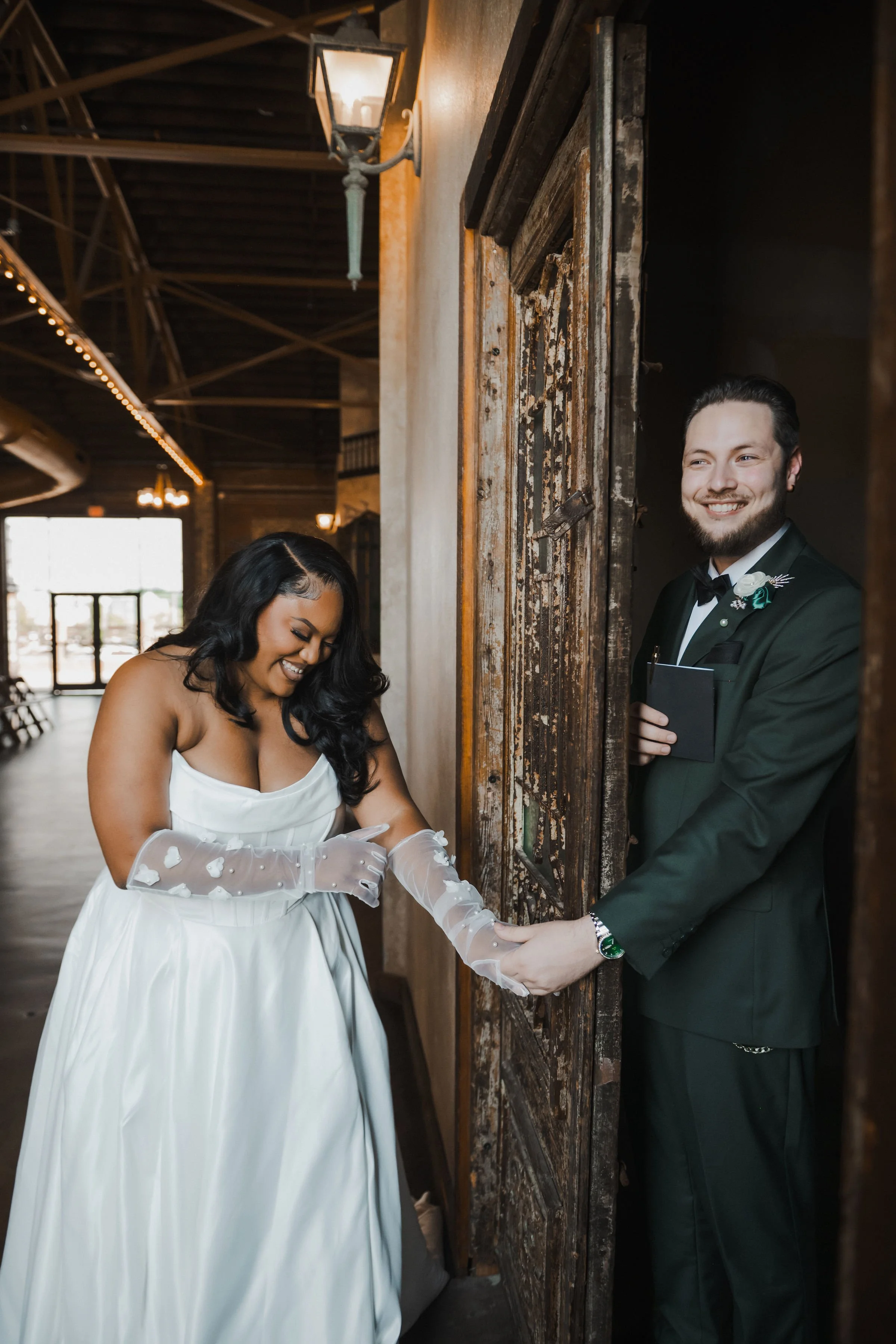 A bride and groom on their wedding day. The bride wearing a white gown and sheer gloves, smiling happily as she touches the groom's hand. The groom in a black tuxedo with a boutonniere, smiling and holding a small notebook, peeking out from behind an