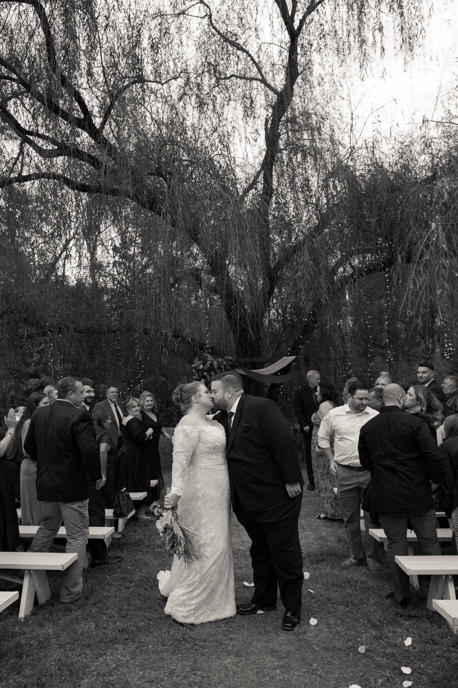 Bride and groom kissing at outdoor wedding ceremony with guests surrounding them under a large tree.