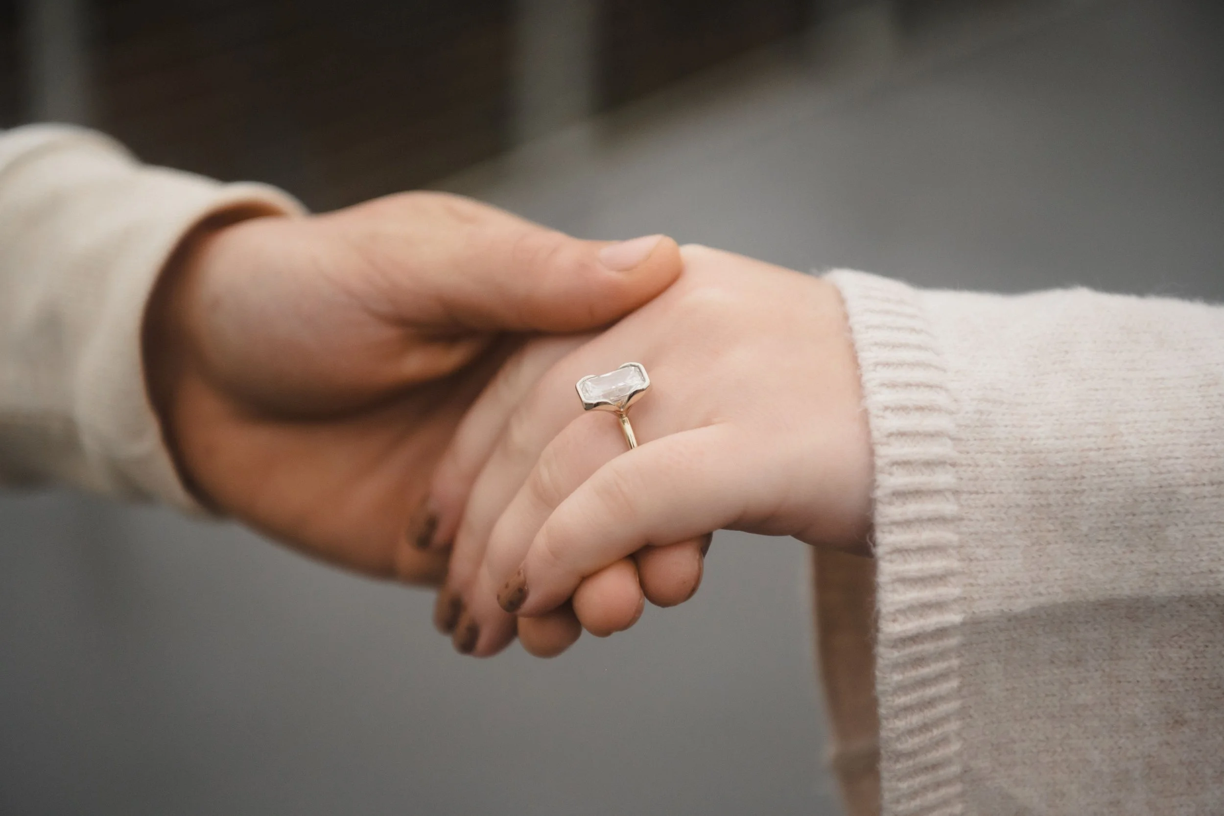 Two hands, one wearing a beige sweater and a ring with a clear, rectangular diamond.