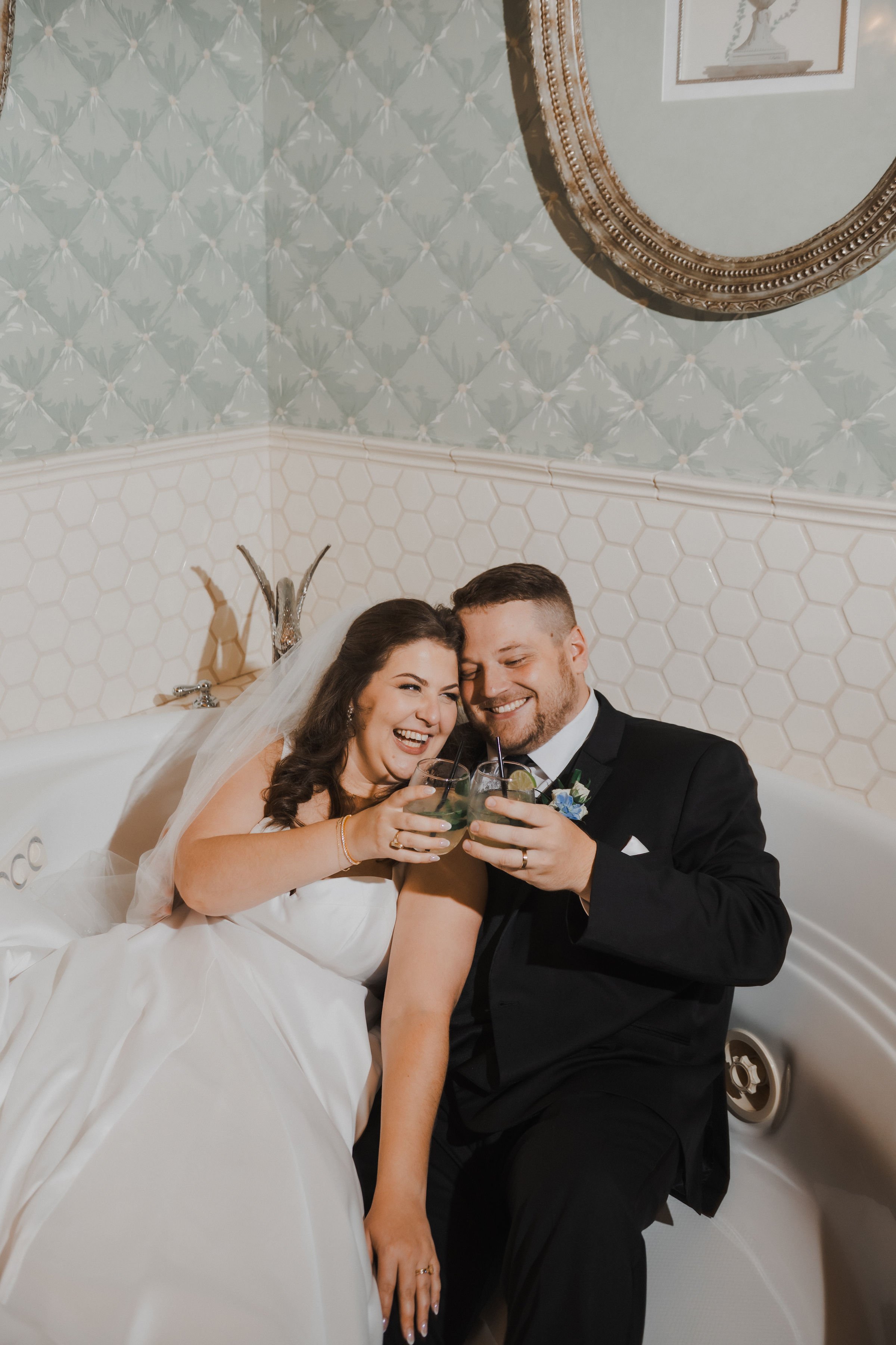 Bride and groom in wedding attire sharing drinks and laughing while sitting in a bathtub.