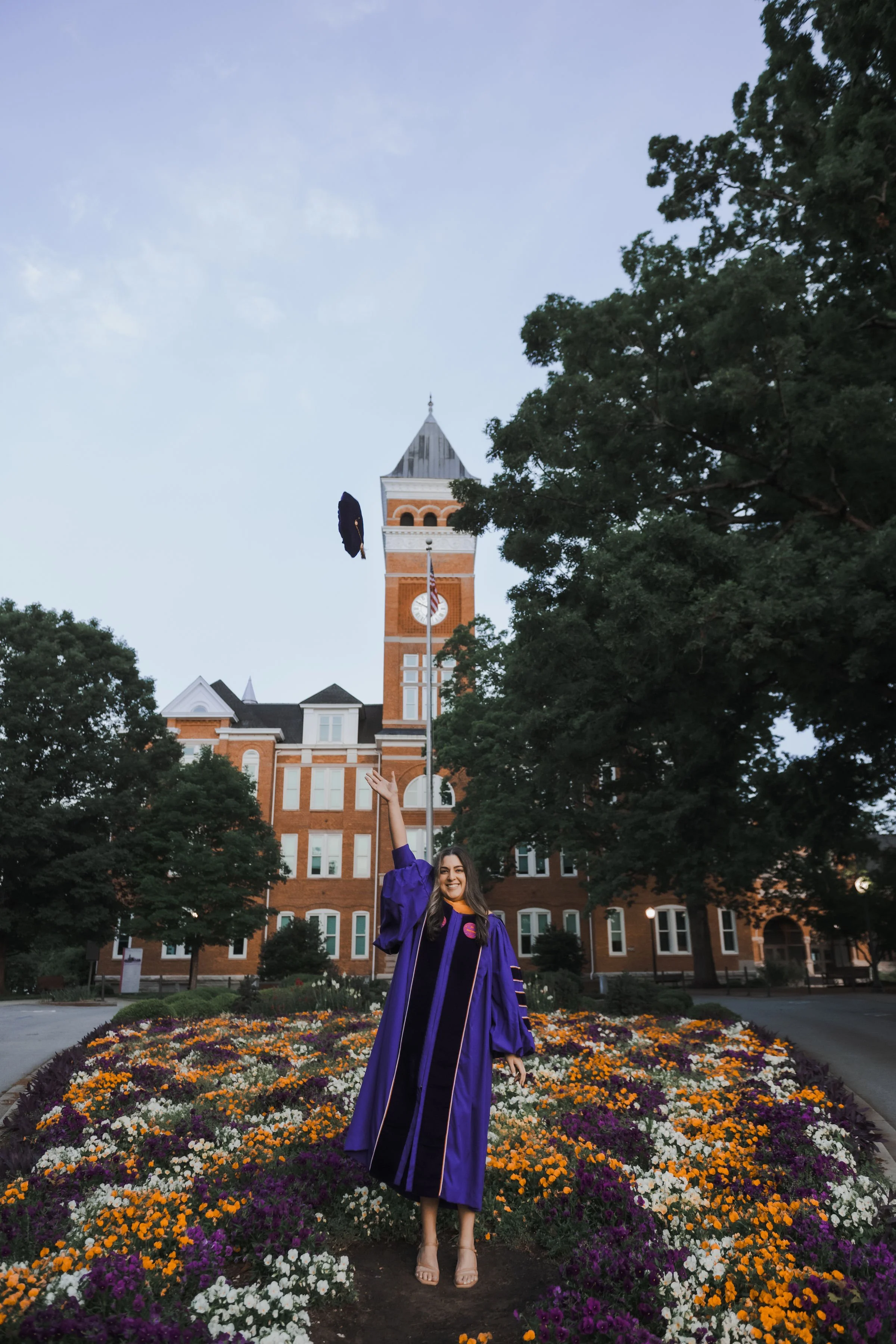 A woman in a purple graduation gown standing on a flower bed, tossing her cap in the air in front of a historic brick building with a clock tower, trees and an American flag.