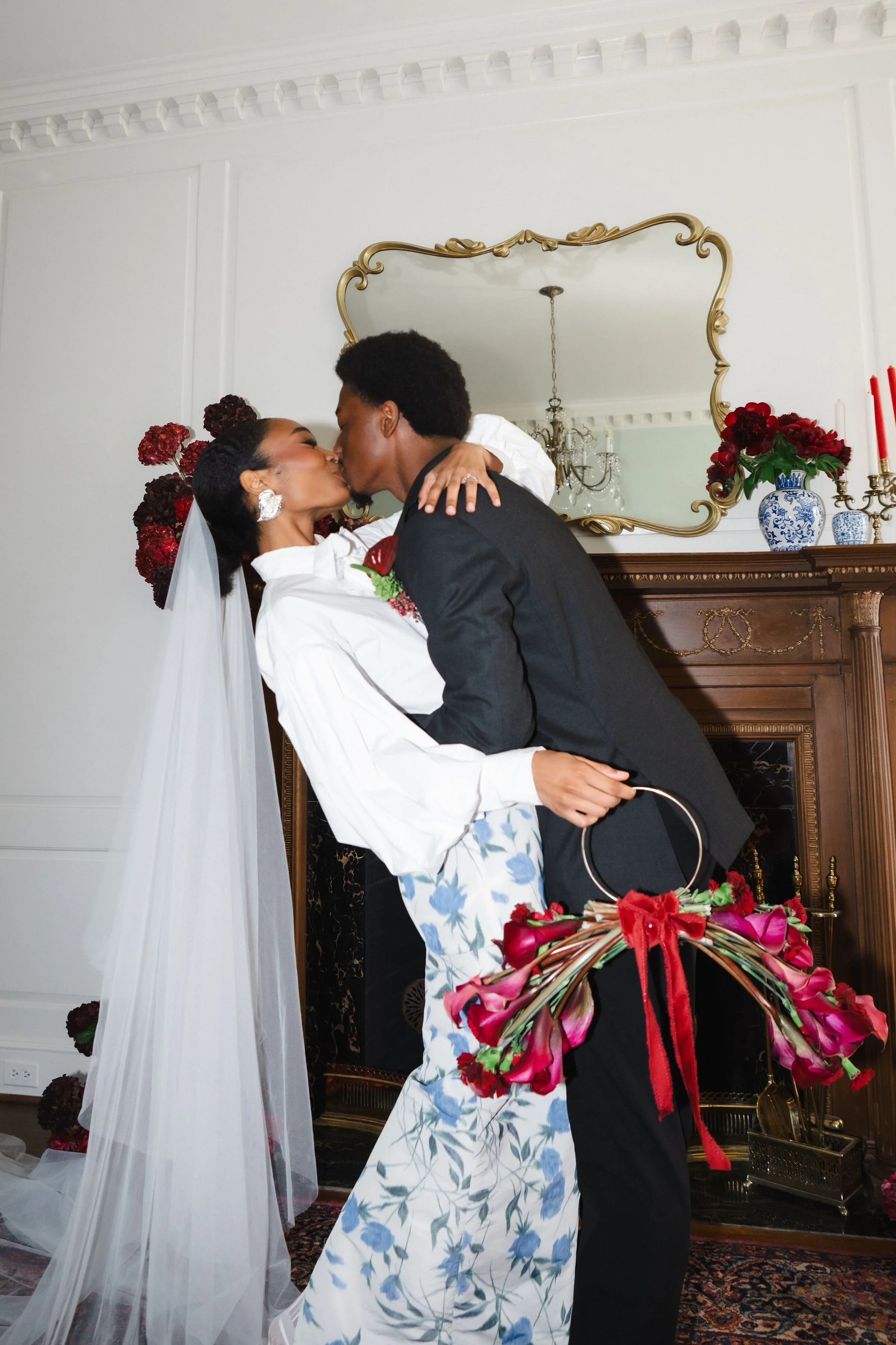 A newlywed couple shares a kiss in a living room decorated with flowers and a mirror above a fireplace.