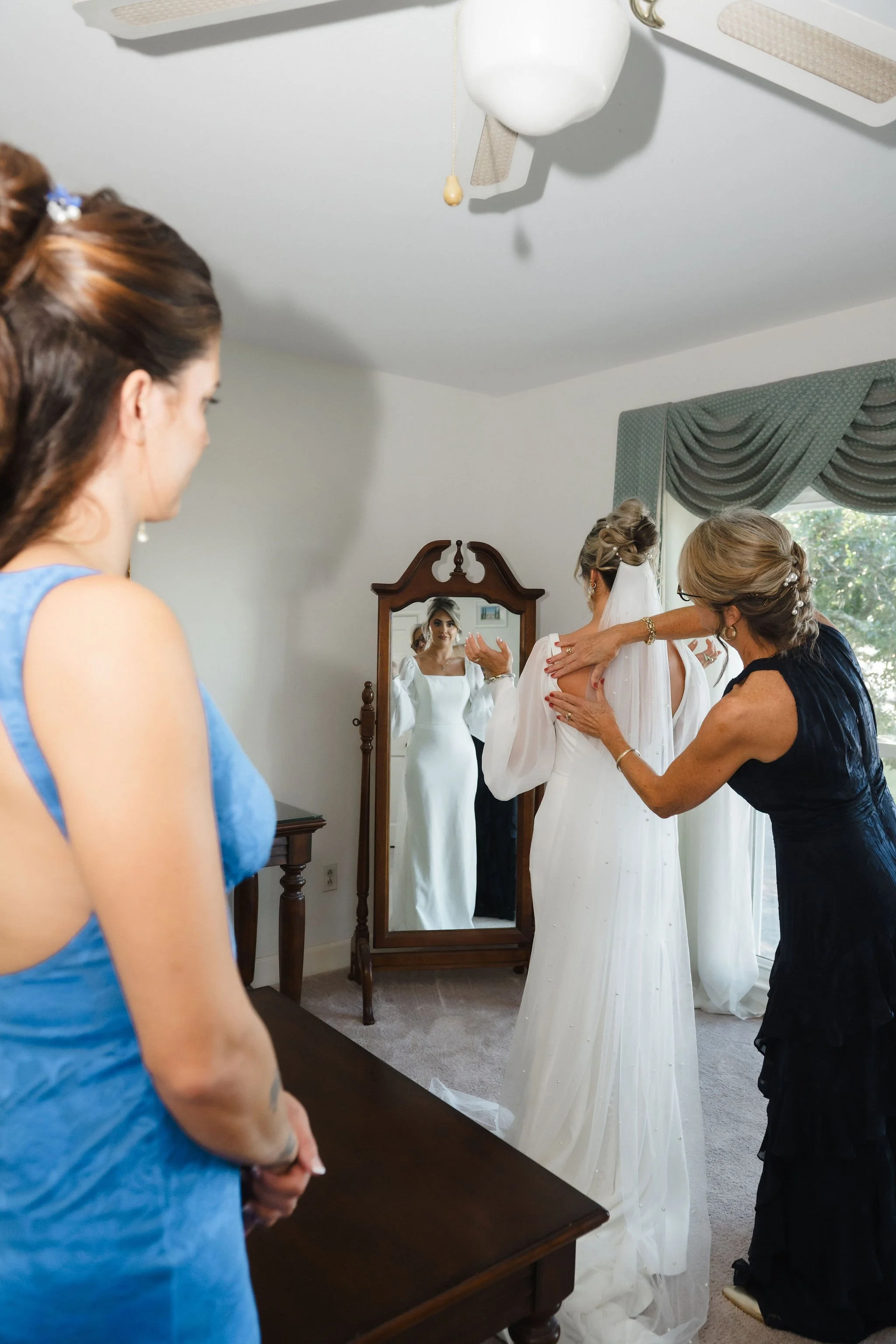 A bride getting help with her wedding dress by her mother and friends in a room, with the bride's reflection visible in a mirror.