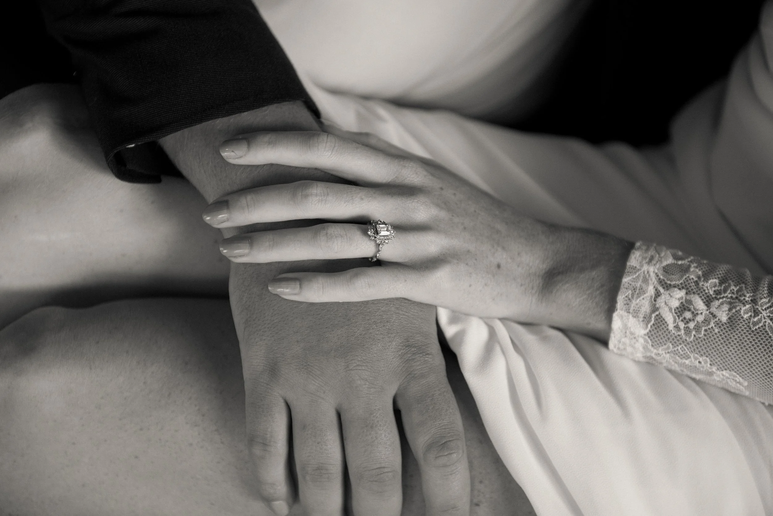 Black and white close-up of a woman’s hand with an engagement ring resting on another woman’s shoulder, with lace sleeve visible.