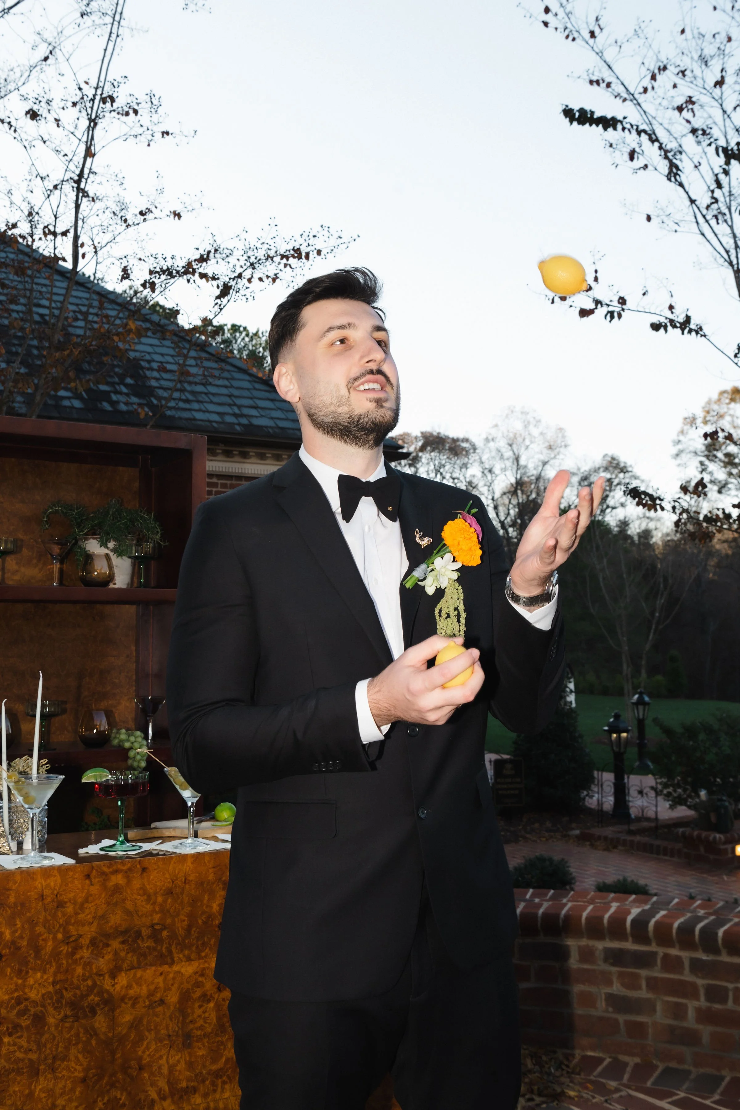 A man in a tuxedo with a flower boutonniere is outdoors, tossing a lemon in the air during evening, with trees and a brick wall in the background.