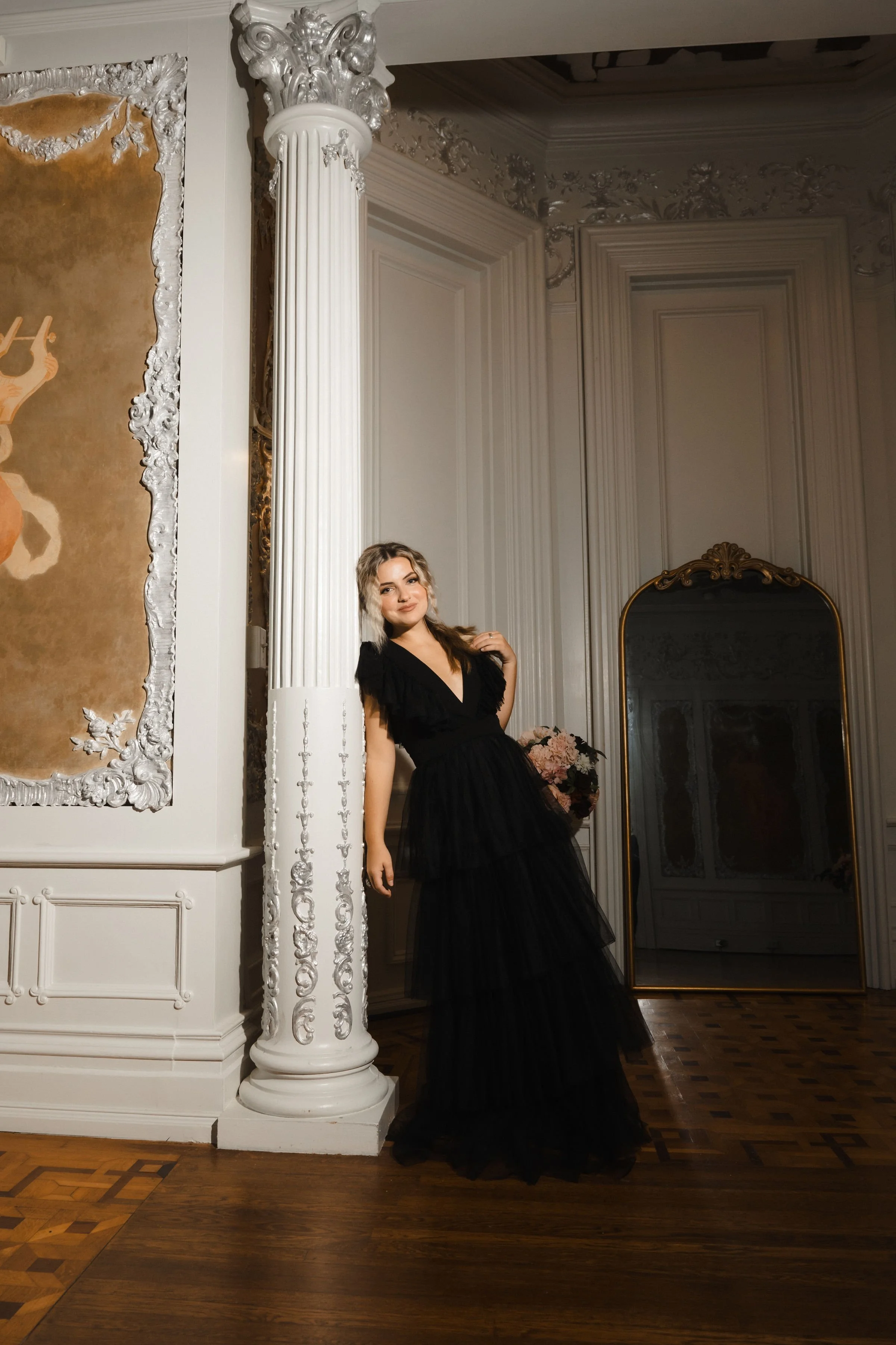 A young woman in a black dress standing next to a white ornate column in a luxurious room with intricate wall moldings and a large mirror.